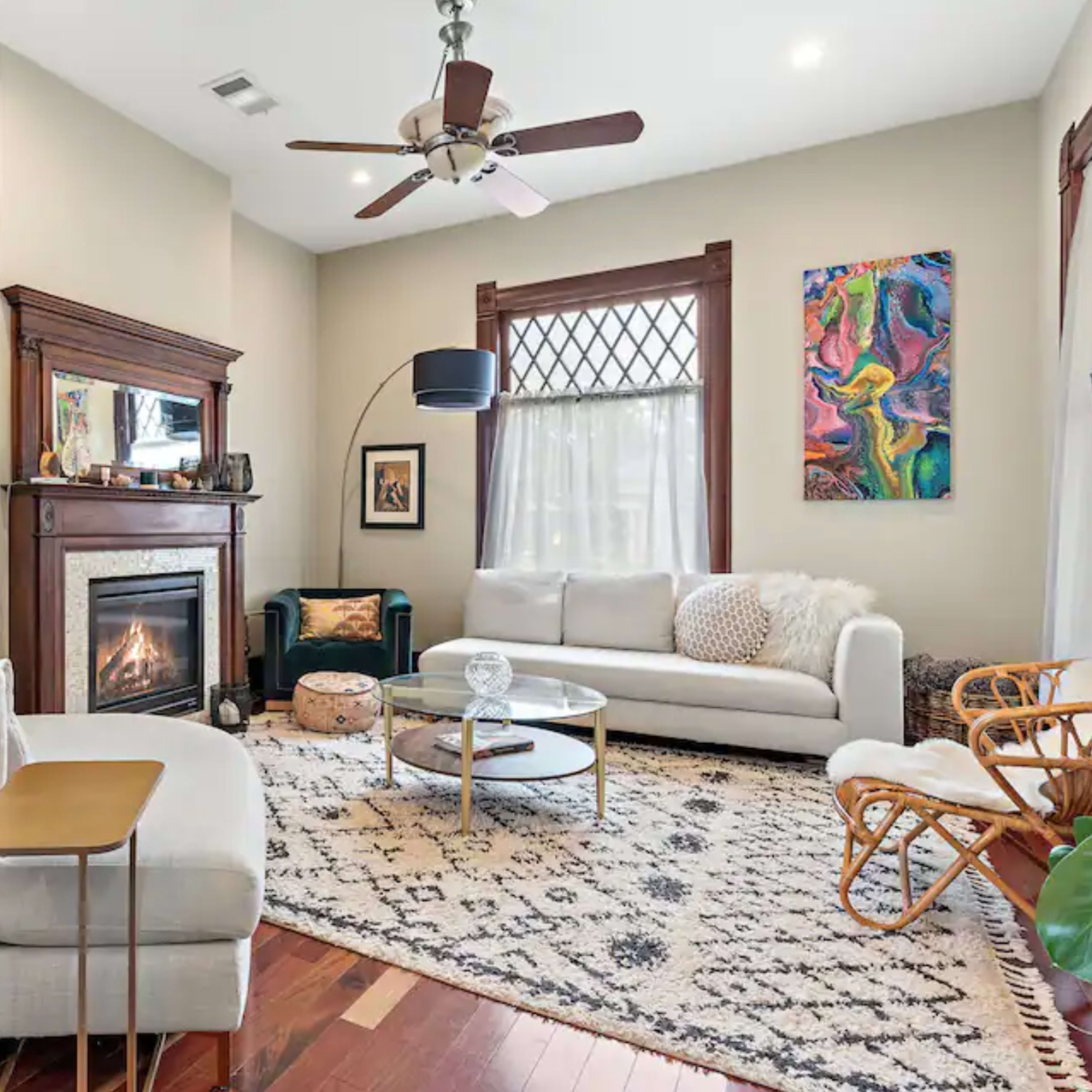 A light-filled living room features a fireplace, a circular glass coffee table, and a mix of stylish furniture on a patterned rug.