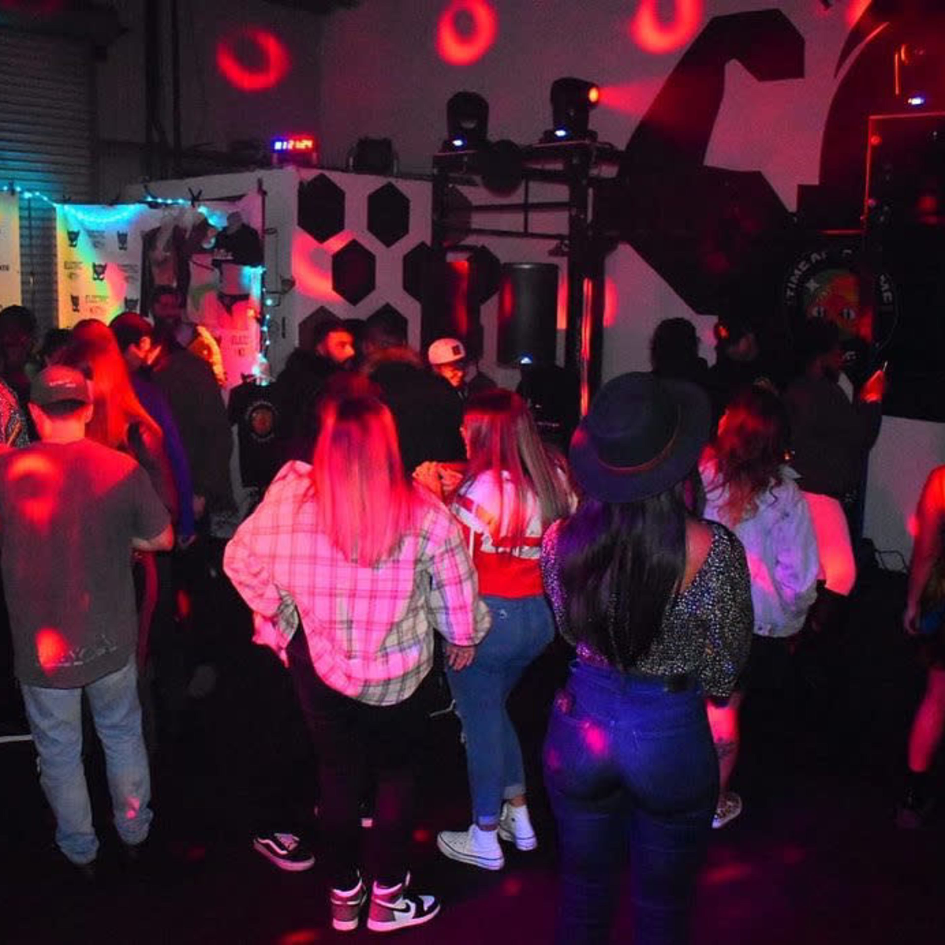 A group of people dance in a dimly lit venue with colorful lights and a DJ setup in the background.
