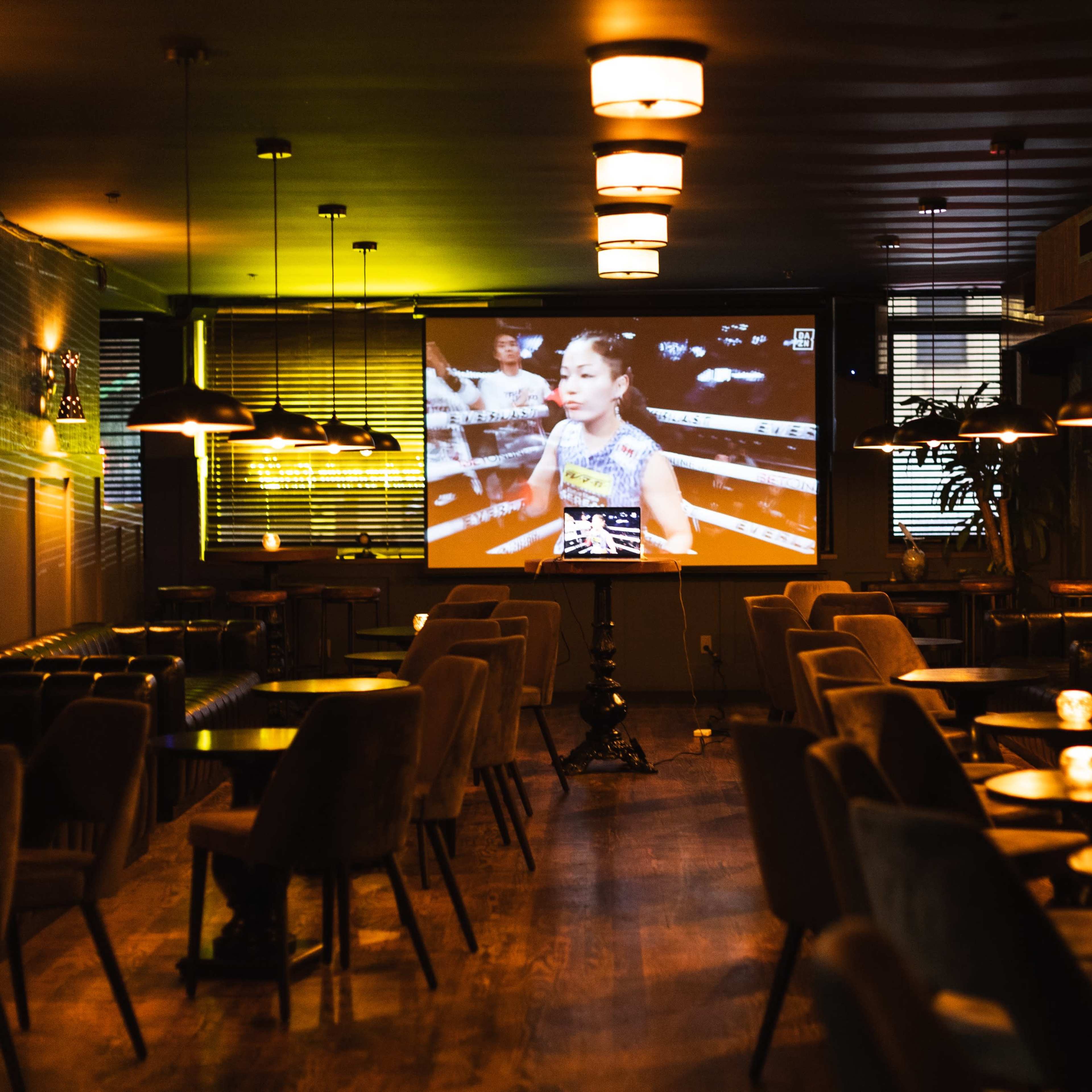 A nearly empty lounge features a large screen displaying a boxing match, with dim lighting and scattered tables.