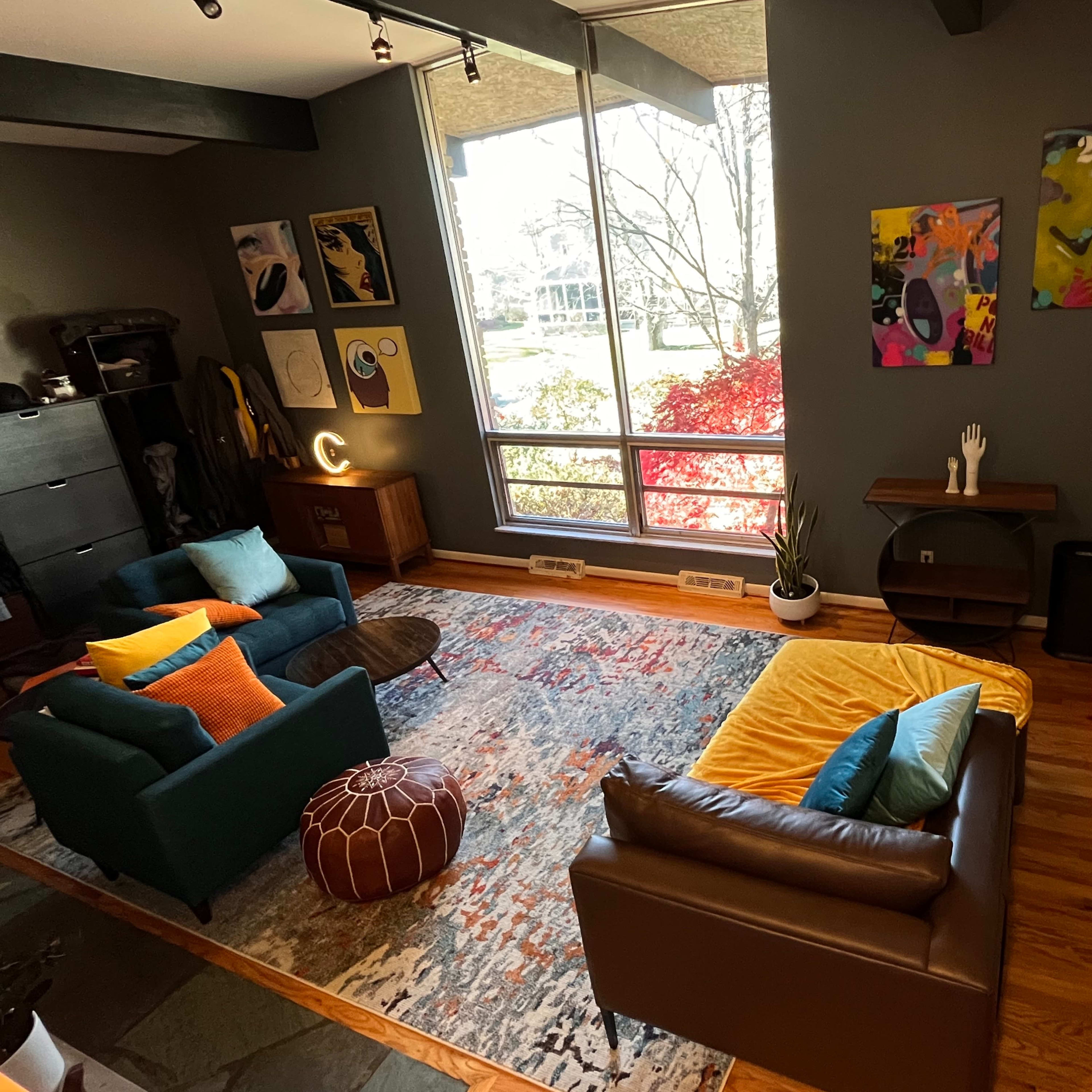 A modern living room with a combination of blue and brown sofas, a colorful rug, and large windows overlooking a garden.