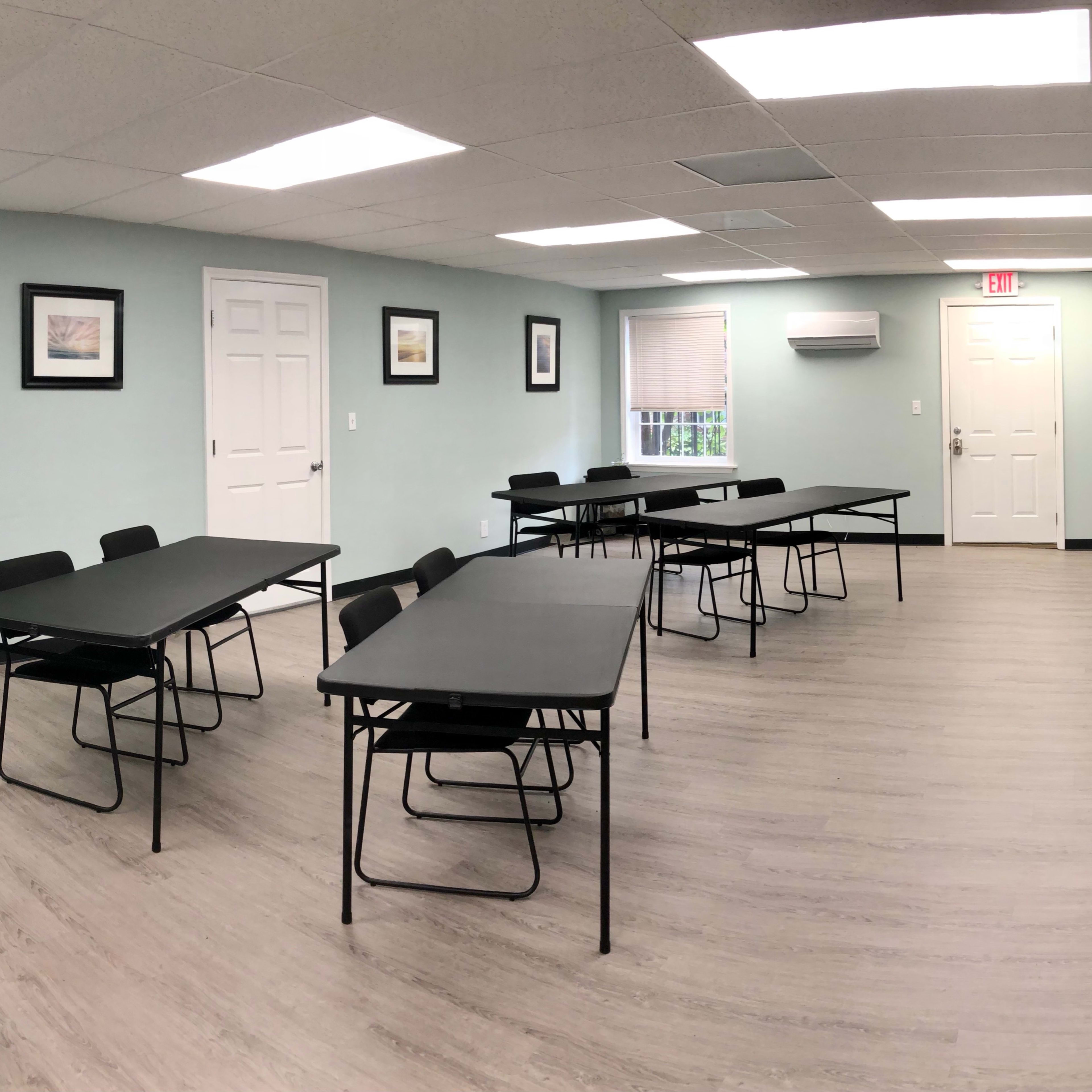 A meeting room with several tables and chairs arranged throughout, featuring light green walls and a wall-mounted television.