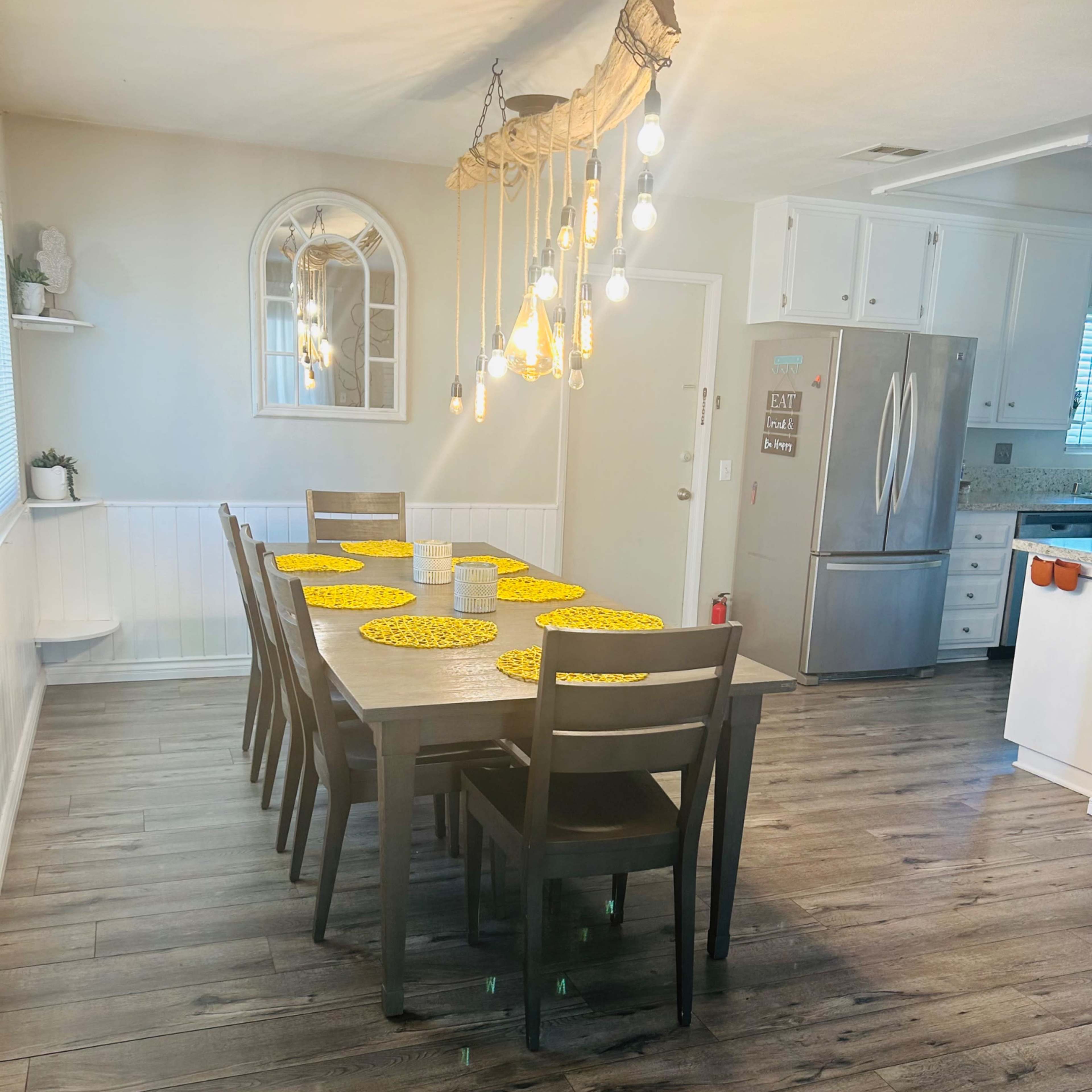 A dining area with a long wooden table set for eight, featuring yellow place mats, under a modern light fixture with hanging bulbs.