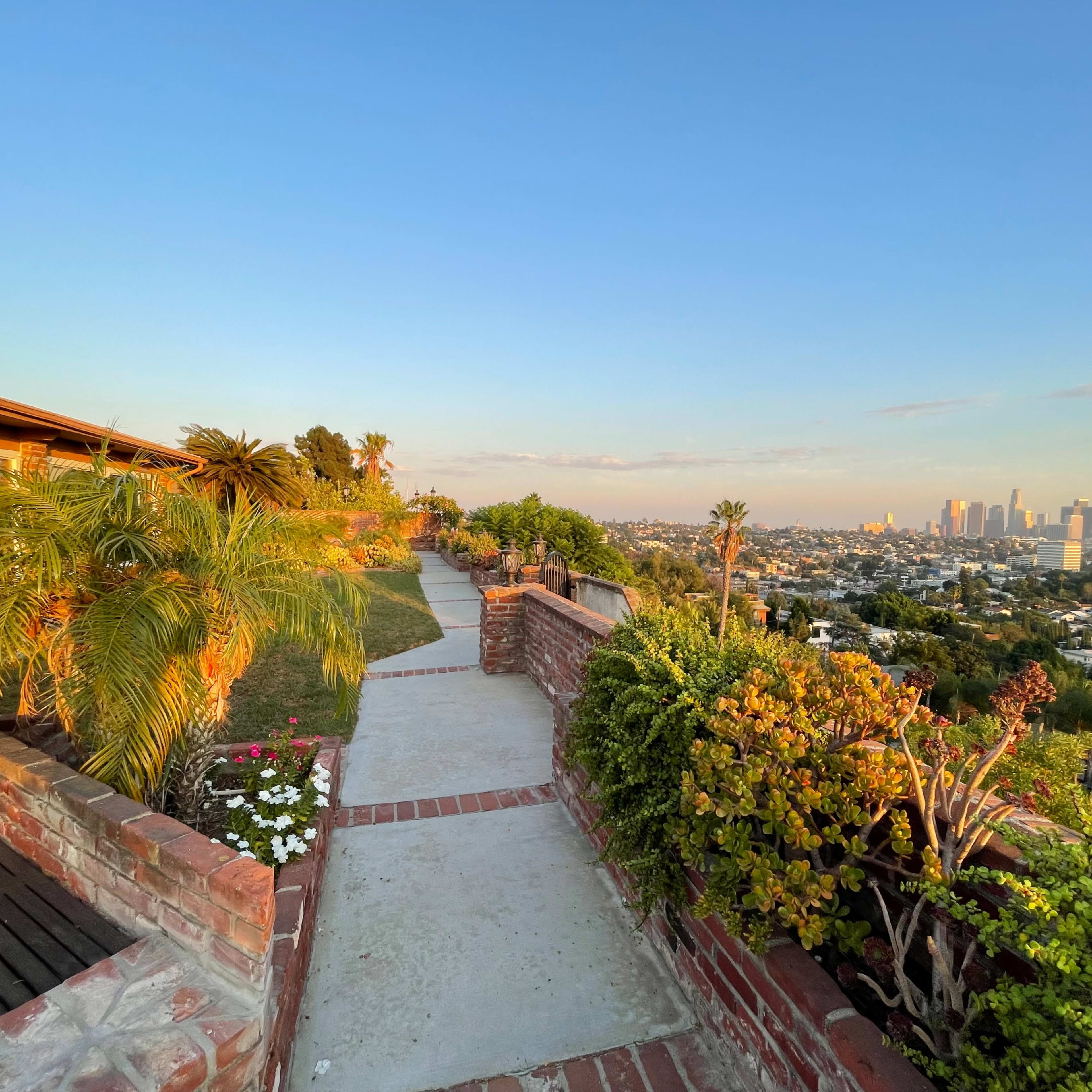 A paved path winds through landscaped greenery, leading towards a view of the city skyline in the distance.
