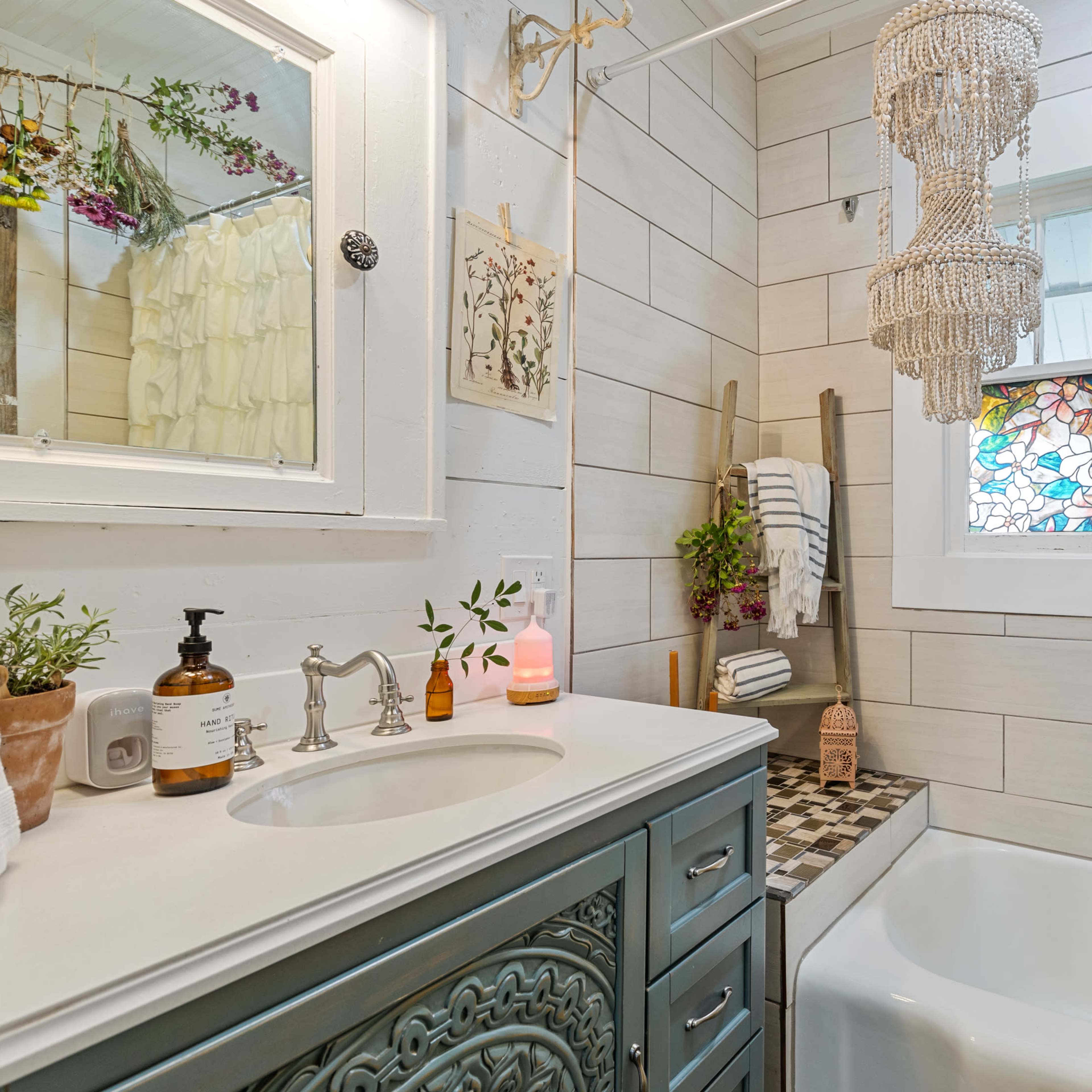 The bathroom features a clawfoot tub, a decorative vanity with a blue finish, and a chandelier above, complemented by a stained glass window and decorative plants.