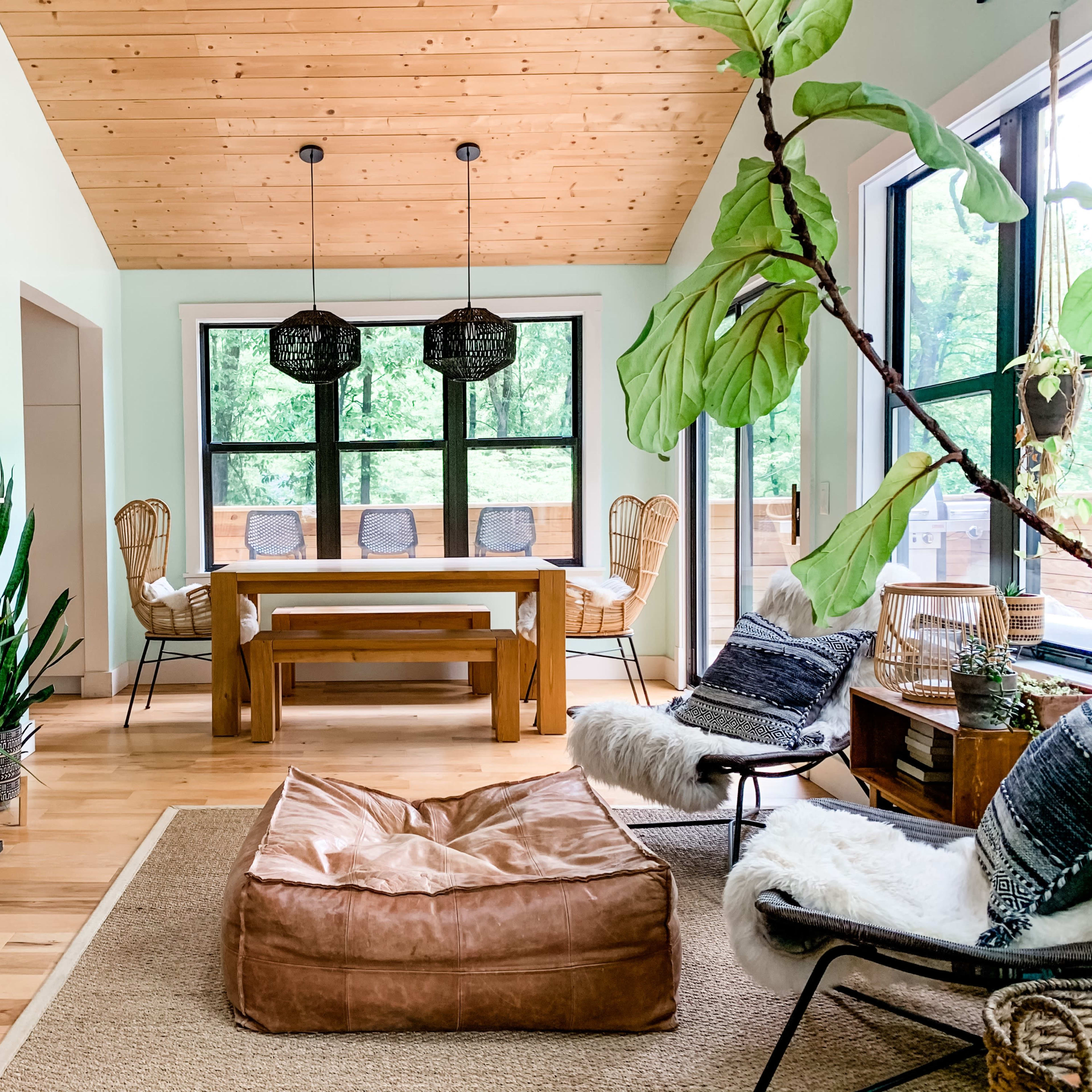 A modern dining area with a wooden table, rattan chairs, a large brown bean bag, and a fireplace, surrounded by greenery and large windows.