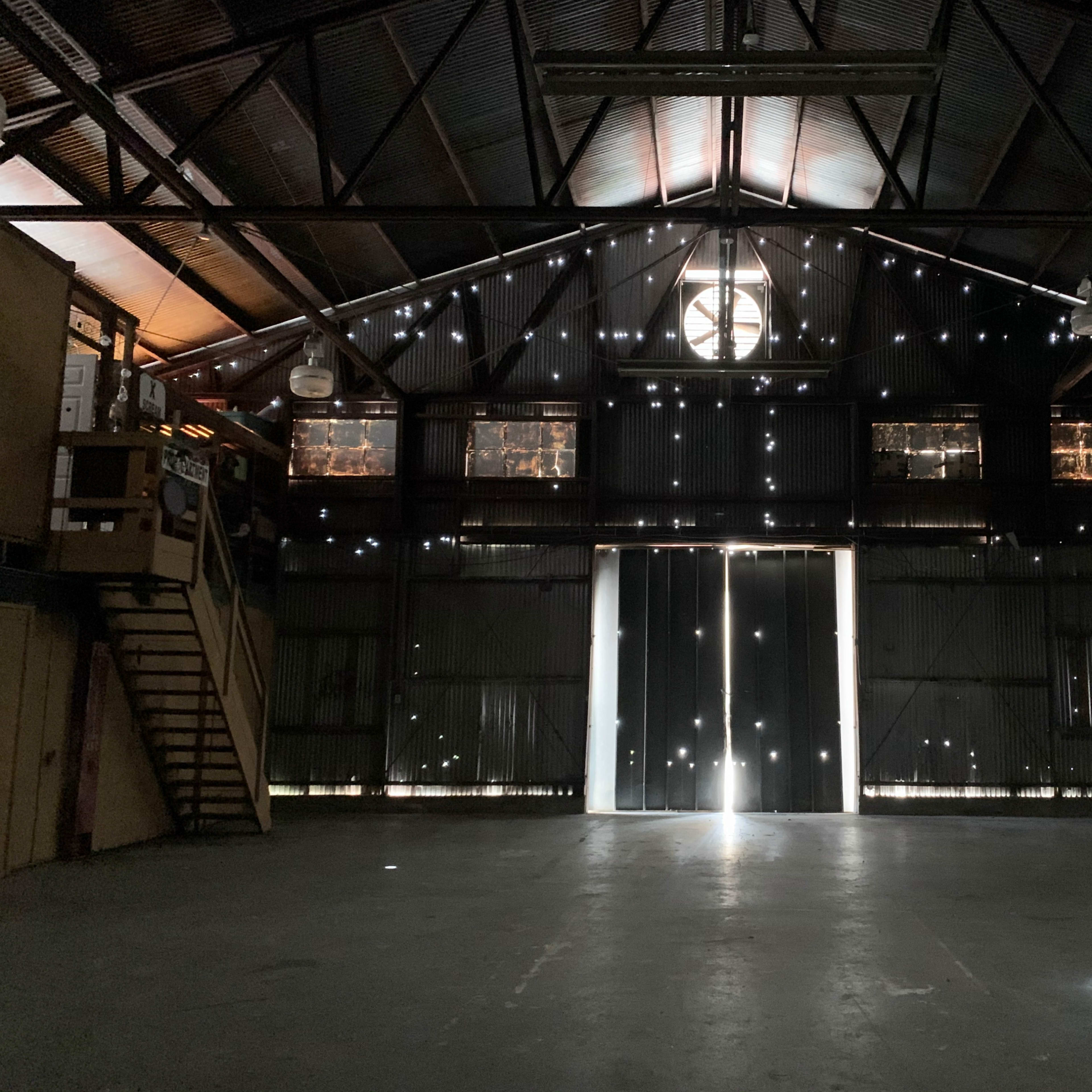 An empty warehouse features large doors at the back, with light streaming in through gaps in the walls and ceiling.