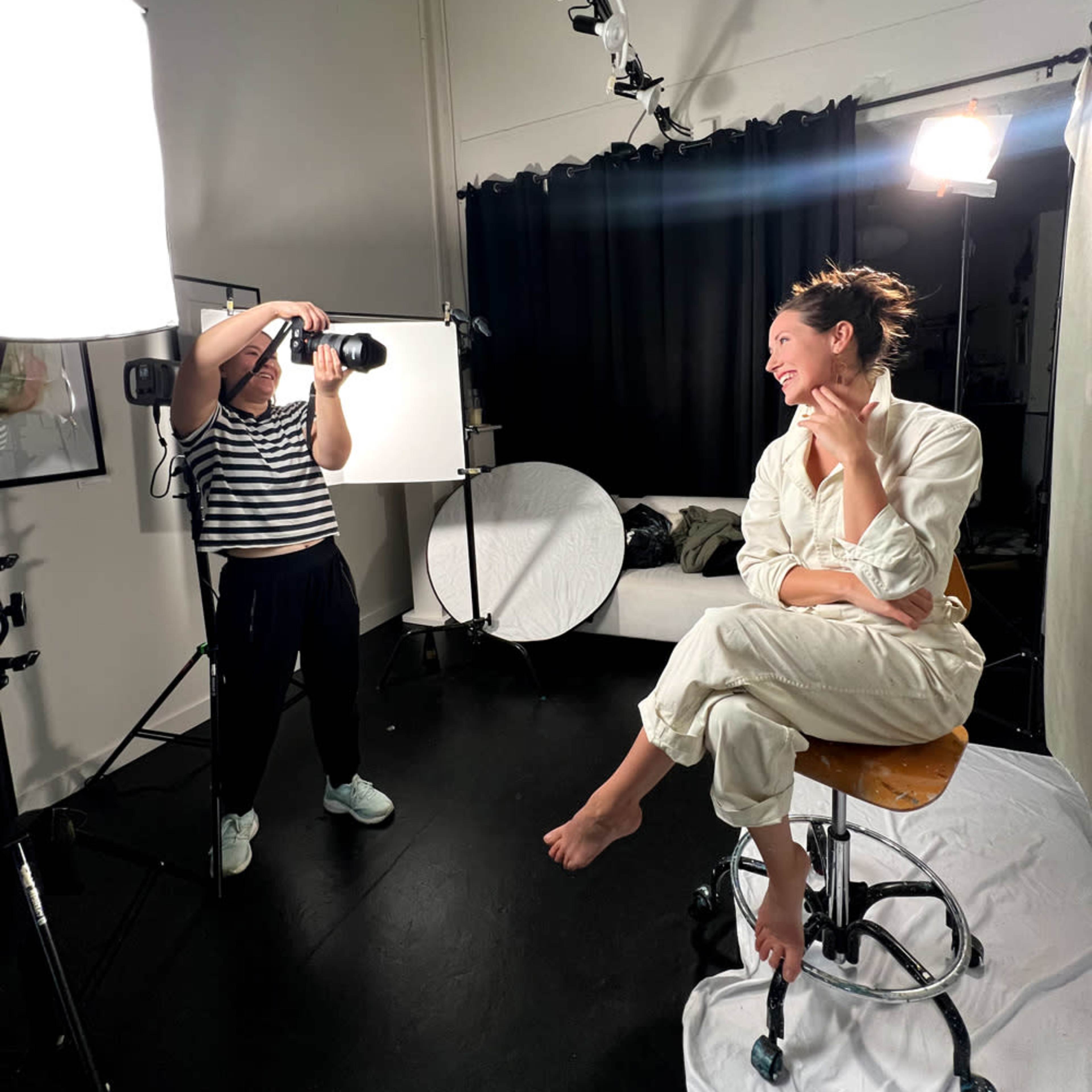 A photographer captures a model seated on a stool in a studio with various lighting equipment and a backdrop.