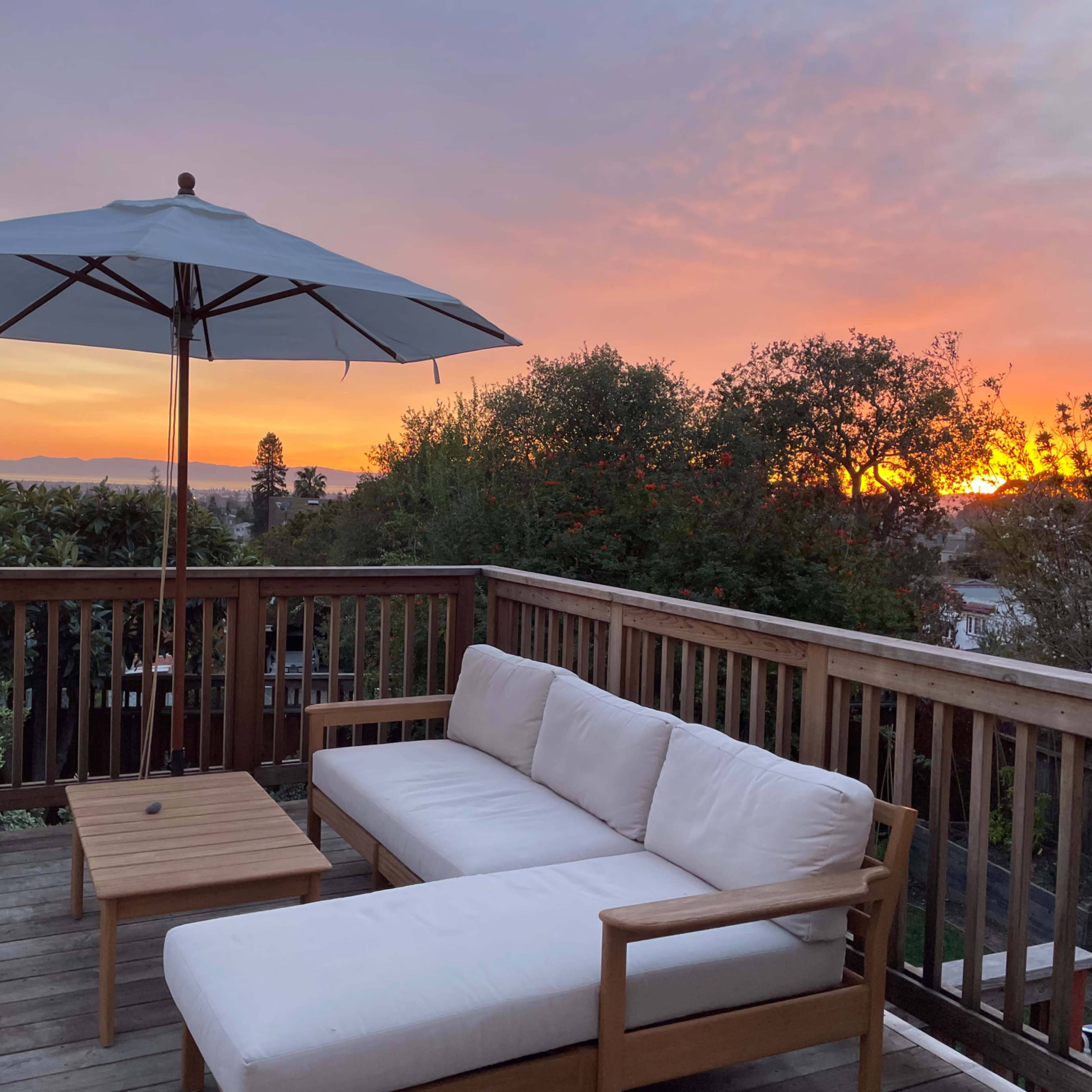A wooden deck with a sofa and table is shaded by a large umbrella, overlooking a sunset with vibrant colors in the sky.