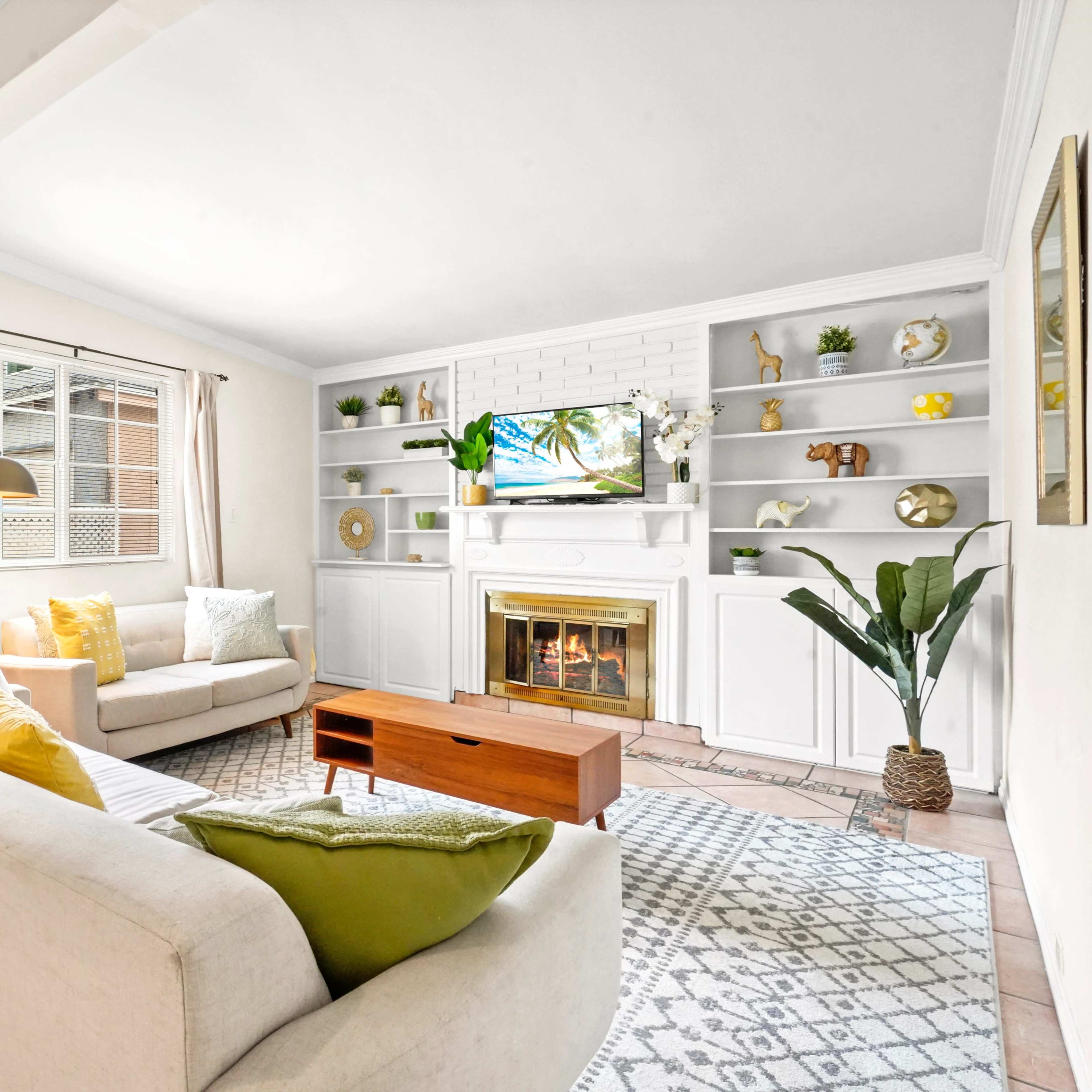 A living room featuring a light-colored sofa, a wooden coffee table, a TV mounted on a white shelving unit, and decorative items on the shelves.