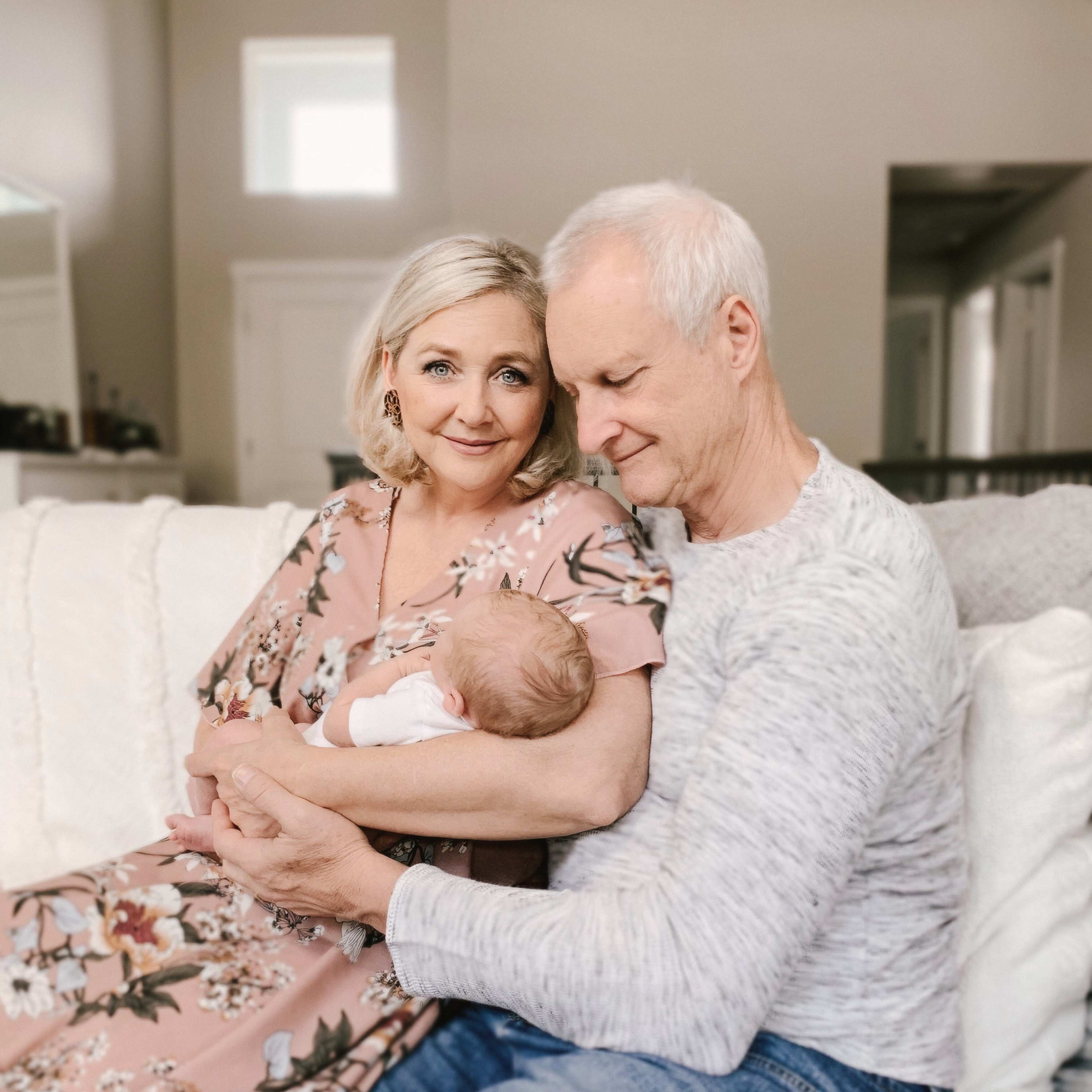 A couple sits together on a couch, holding a newborn baby in their arms, with soft natural light illuminating the room.