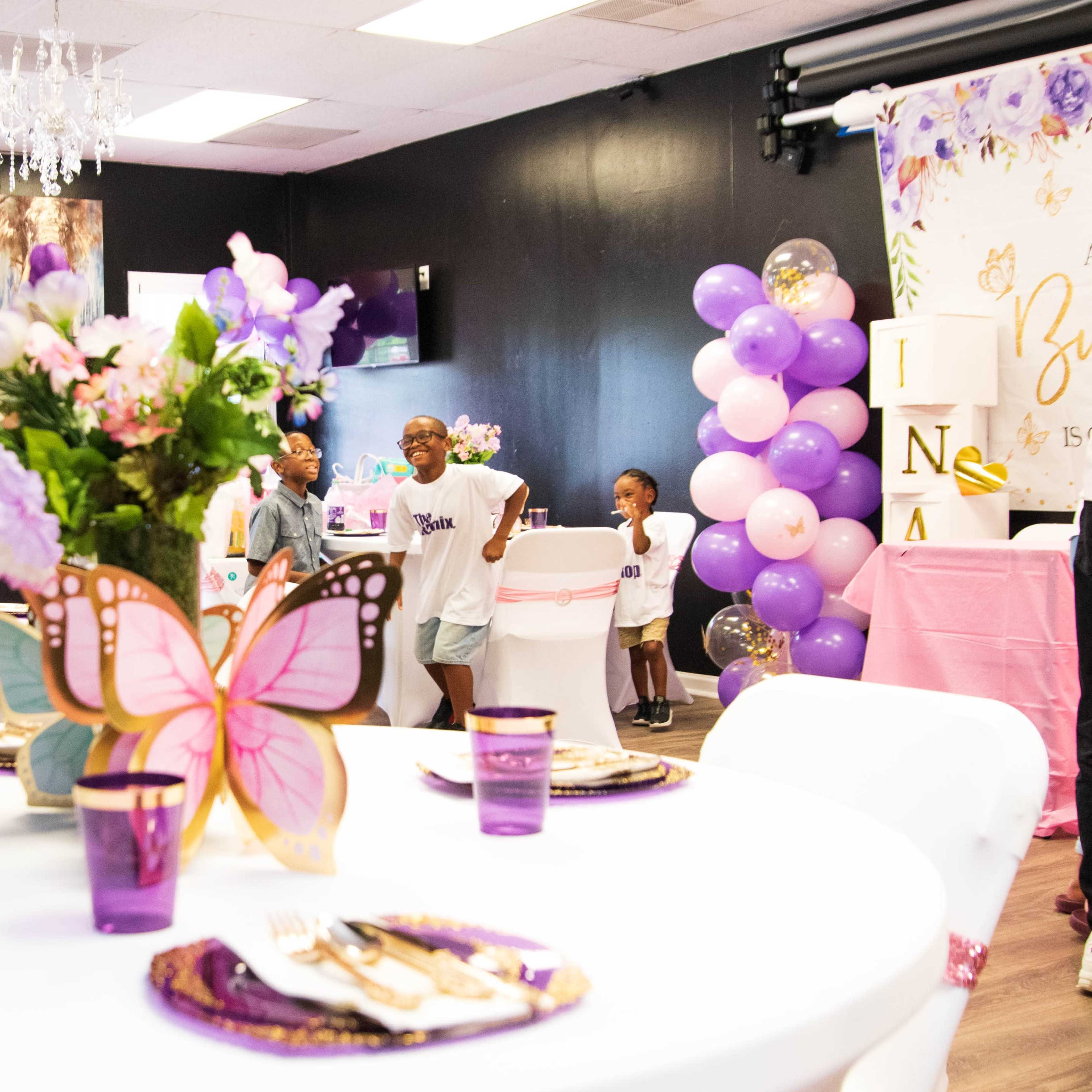 A decorated event space with round tables set up for a celebration, featuring butterfly-shaped centerpieces and purple decorations.