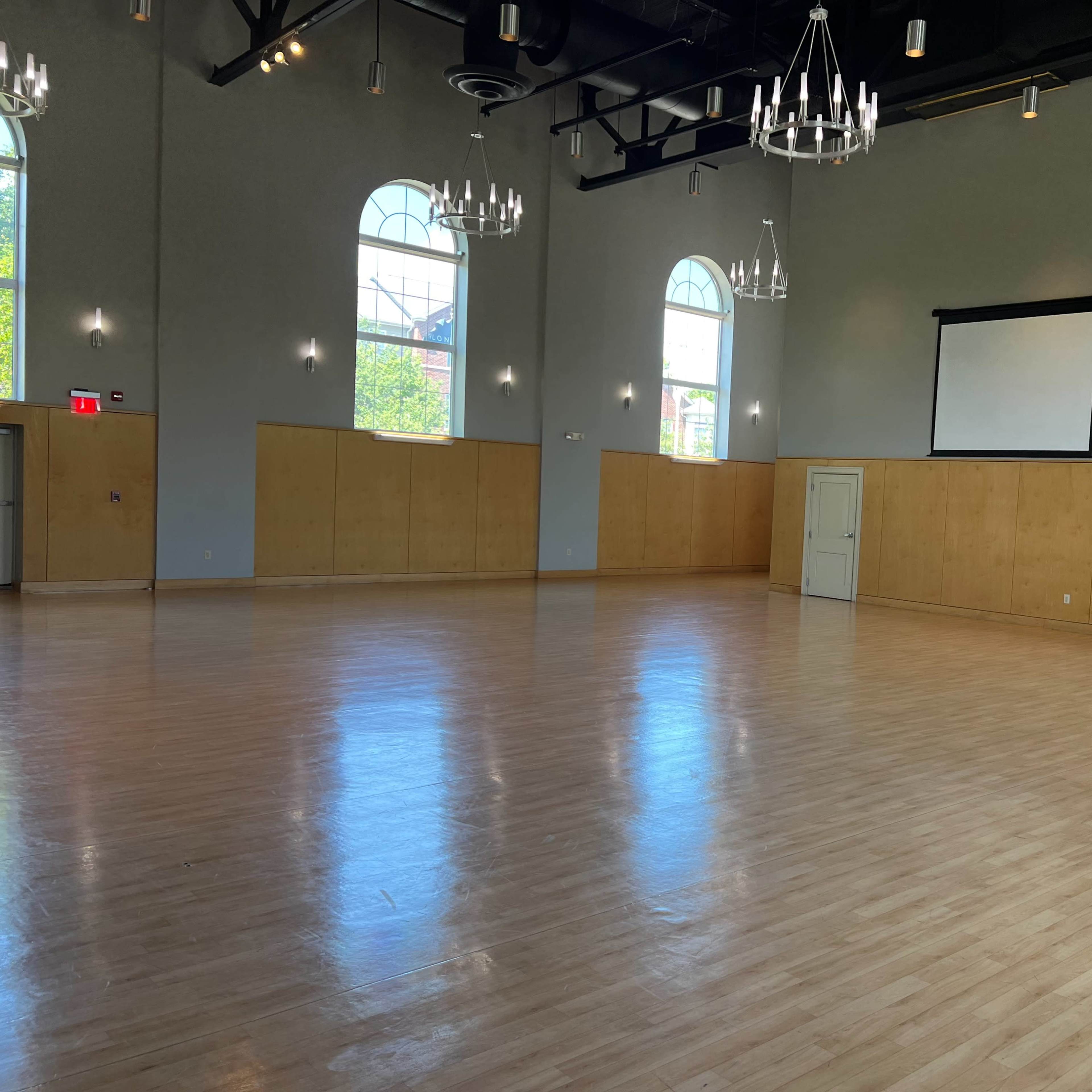 A spacious, empty hall with wooden flooring, large windows, and chandeliers hanging from the ceiling.