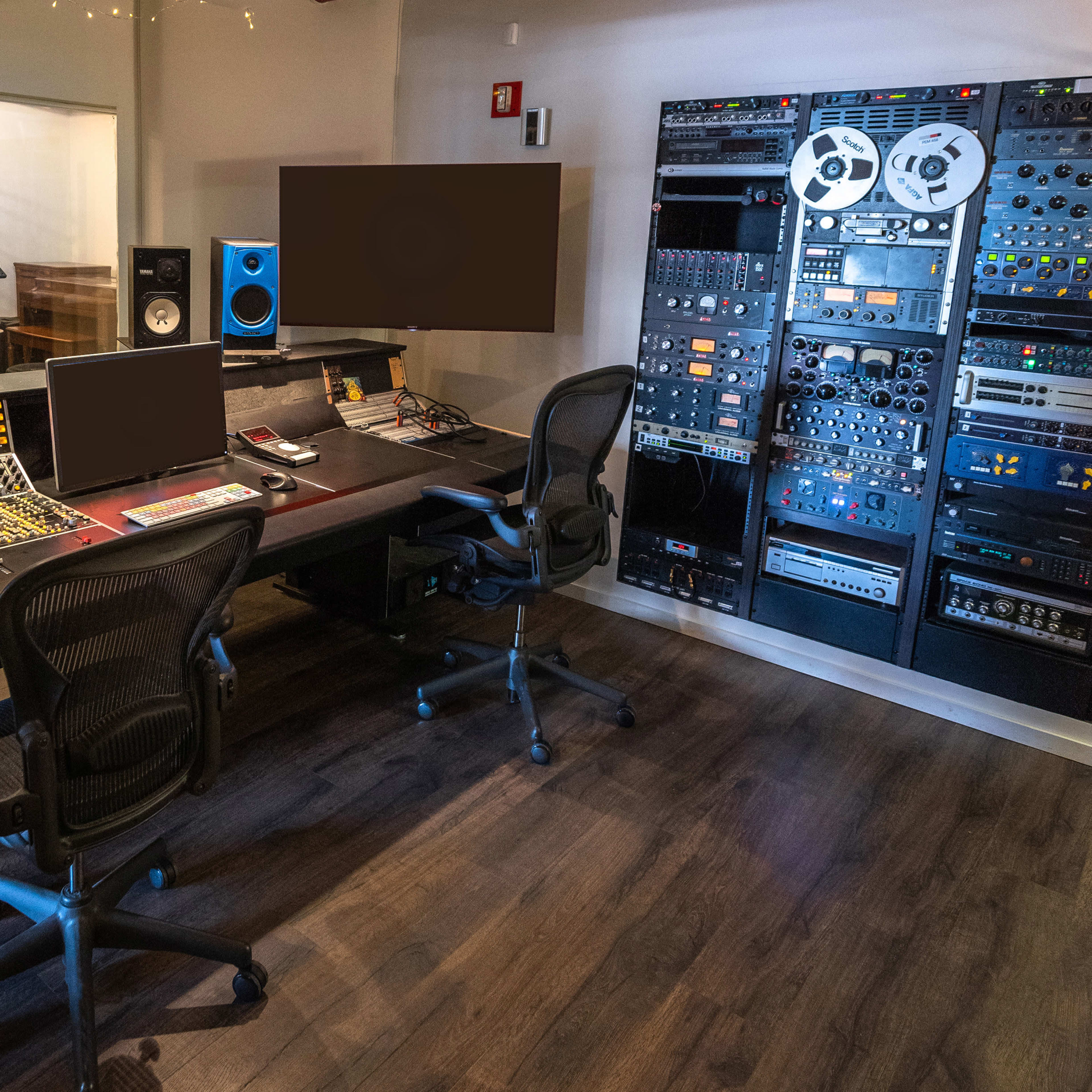 A modern recording studio with a large mixing console, two computers, and various audio equipment mounted on the wall.