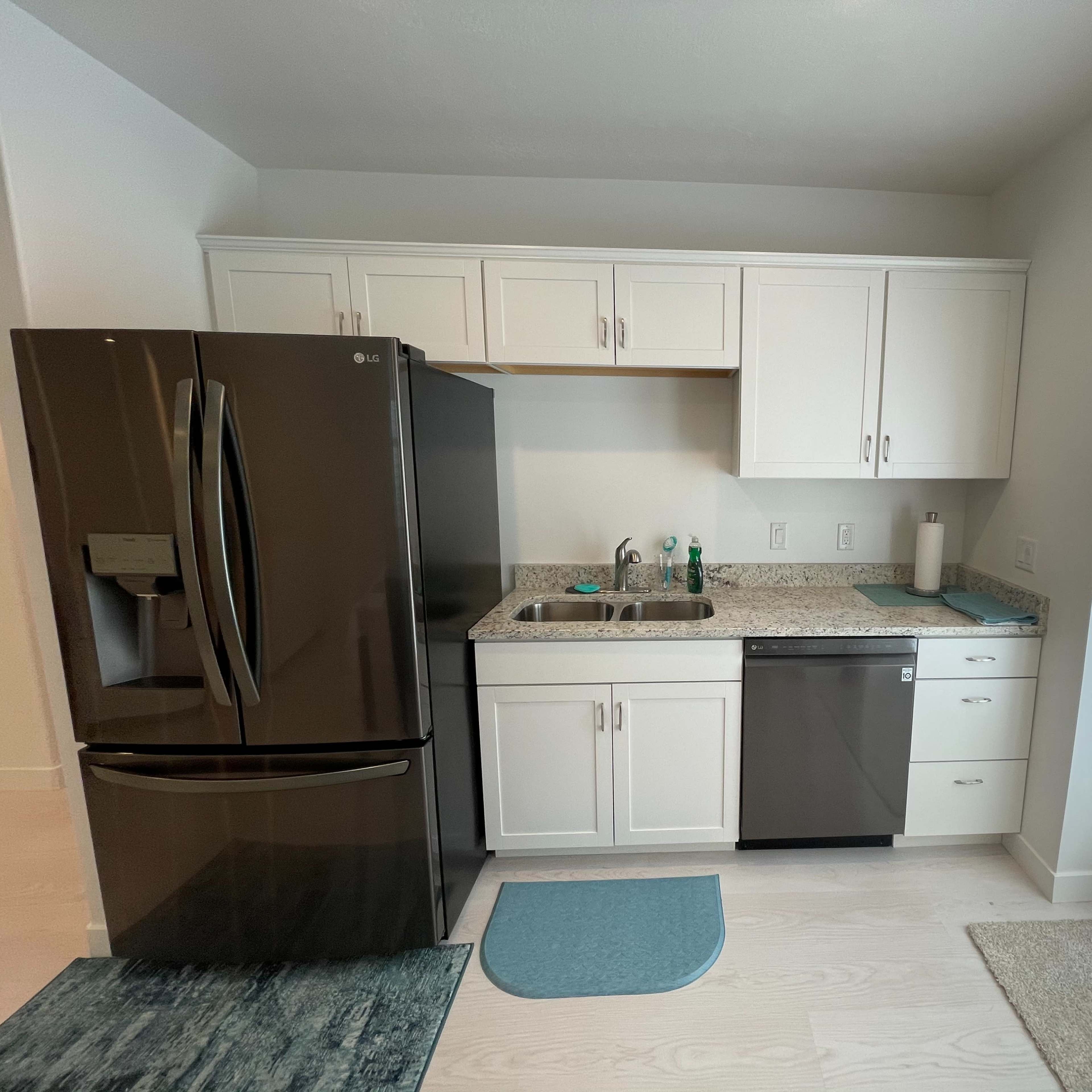 A modern kitchen with white cabinets, a granite countertop, a black refrigerator, a stainless steel dishwasher, and a window with blinds.