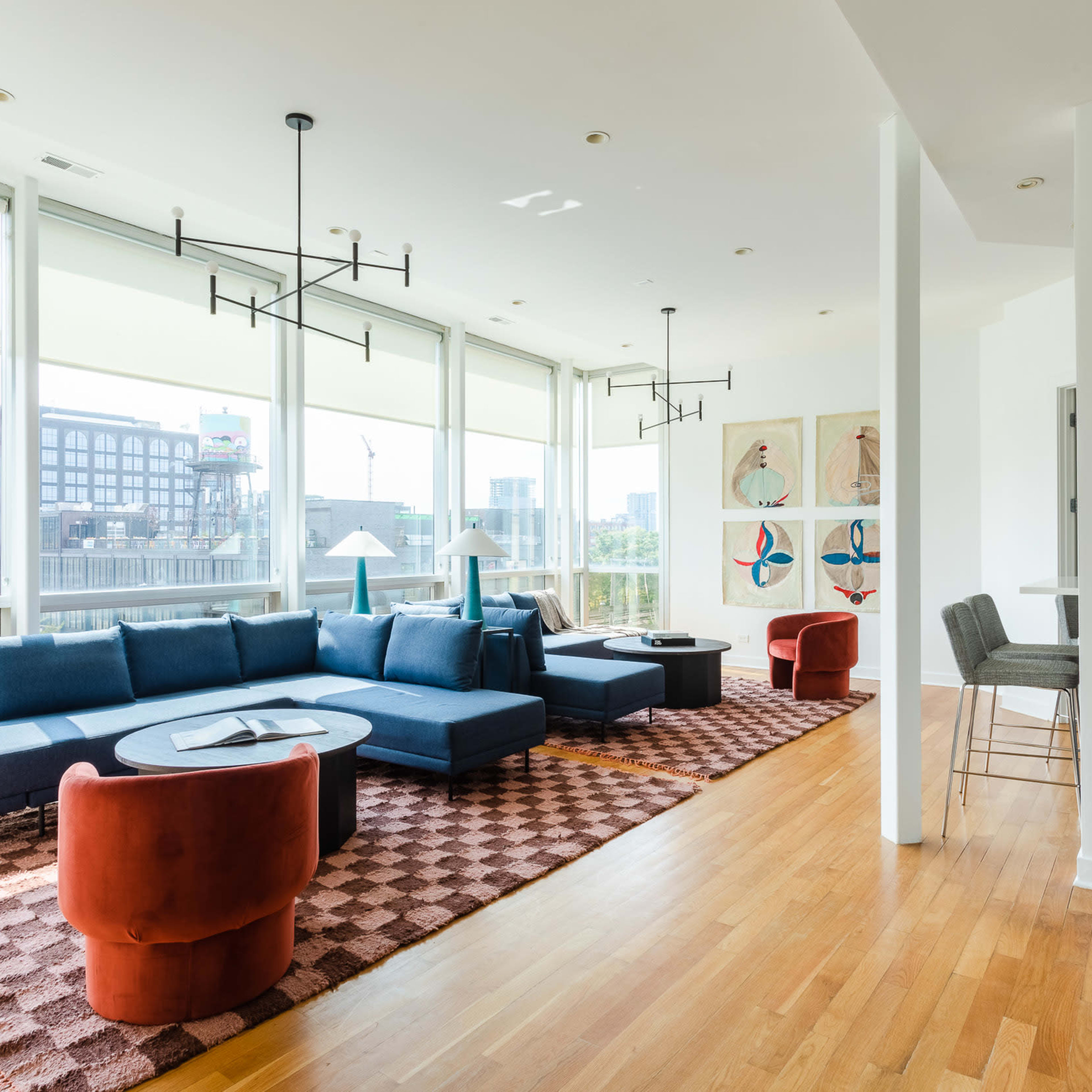 A modern living space featuring a blue sectional sofa, two red accent chairs, and a large area rug, with large windows offering a view of the city.