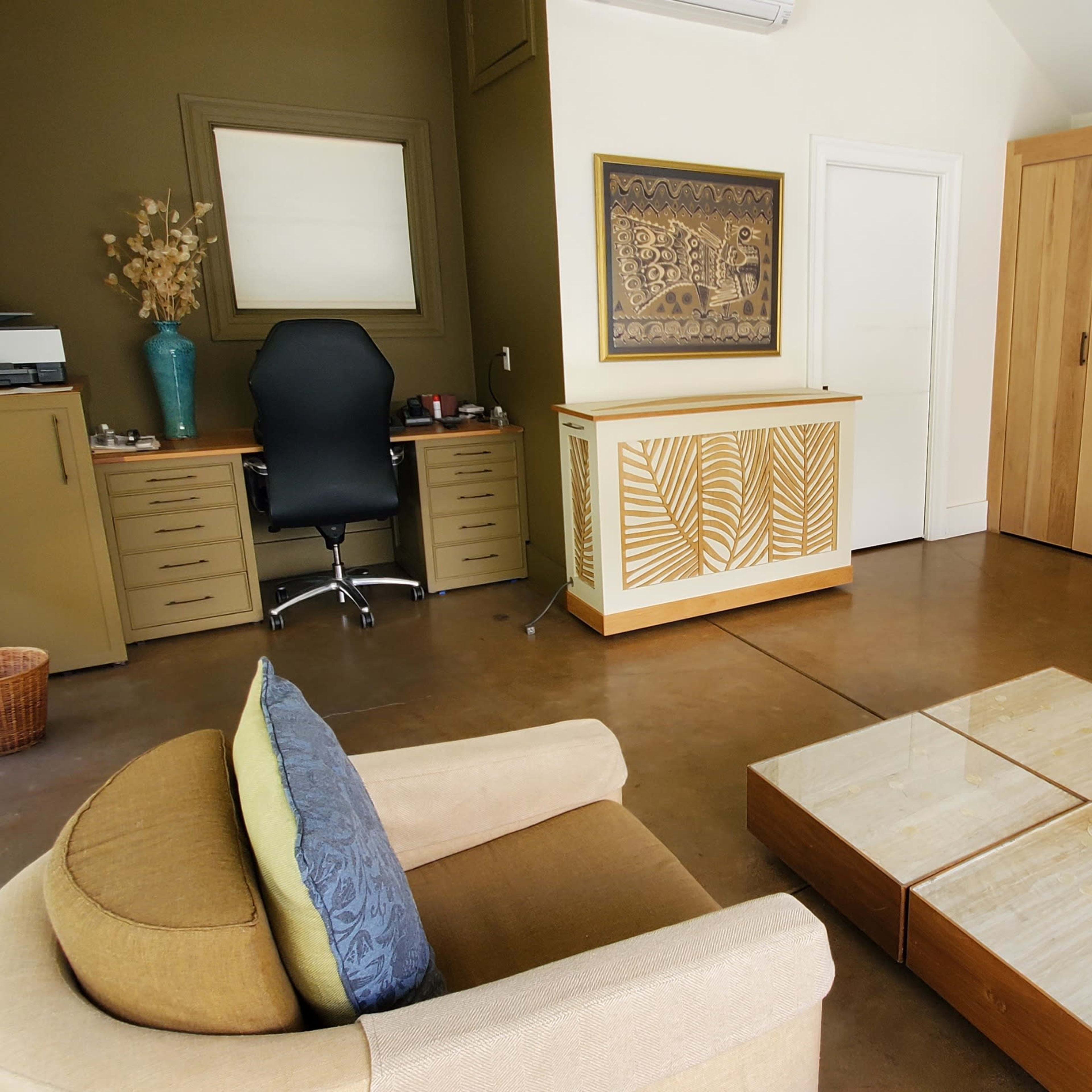 A small office space featuring a desk with a chair, a set of wooden storage cabinets, two armchairs with cushions, and a coffee table in the center.
