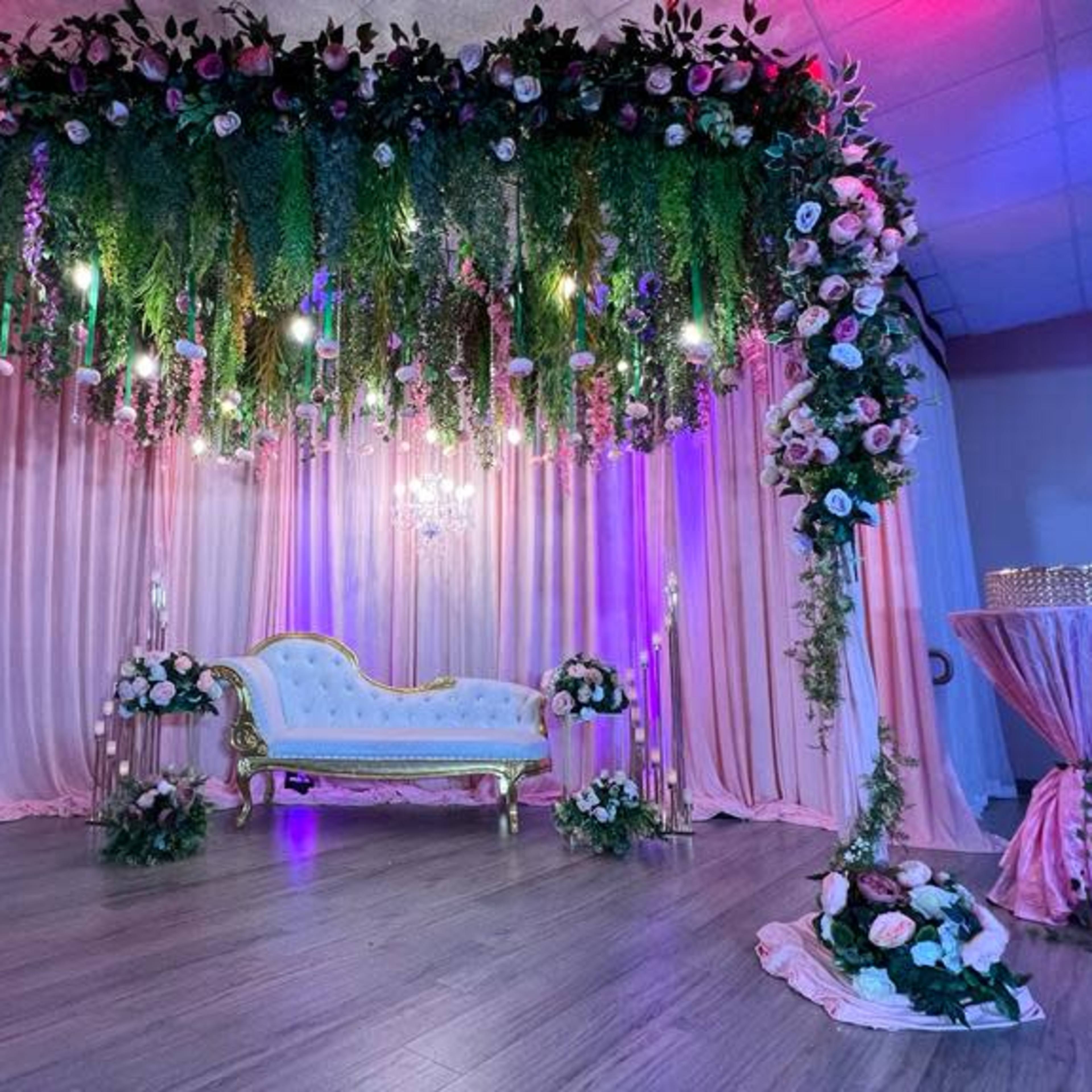 The scene features a beautifully decorated event space with a lavish floral backdrop, a vintage white sofa under hanging greenery and lights, and a pink draped table nearby.