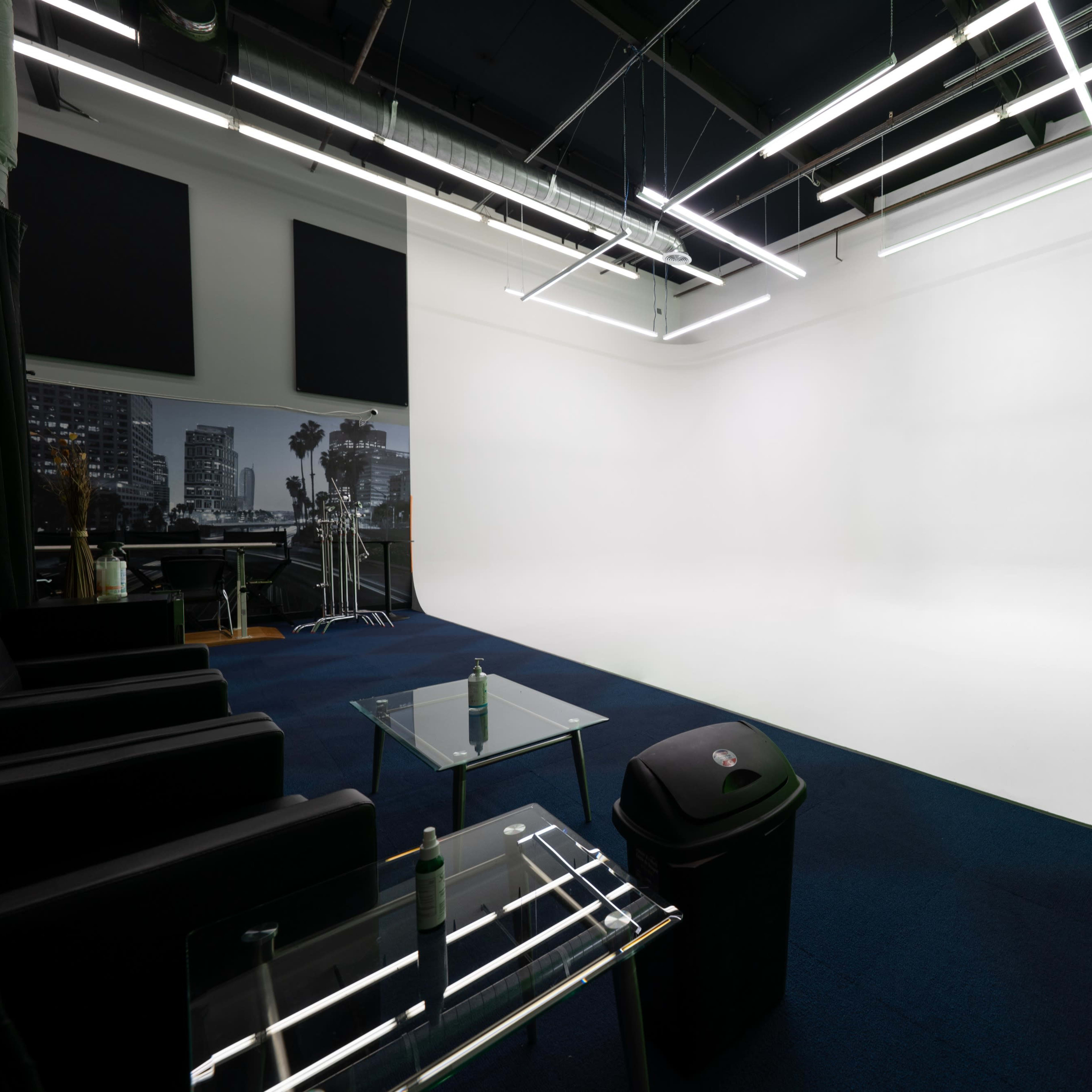 A photography studio featuring a large white backdrop, modern lighting fixtures, and a seating area with glass tables.