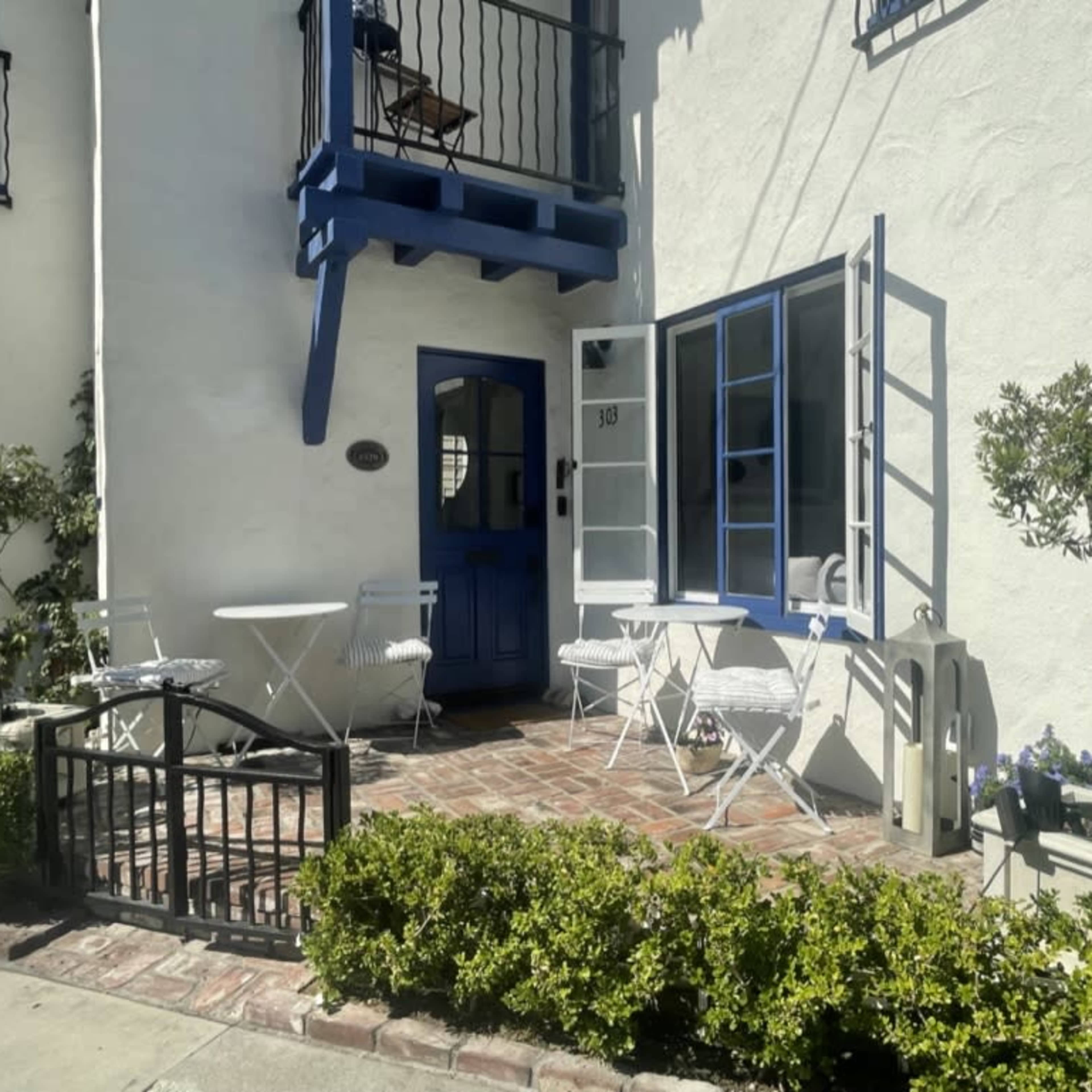 A small outdoor patio features a round table and white chairs, framed by a blue door and window against a white stucco wall.