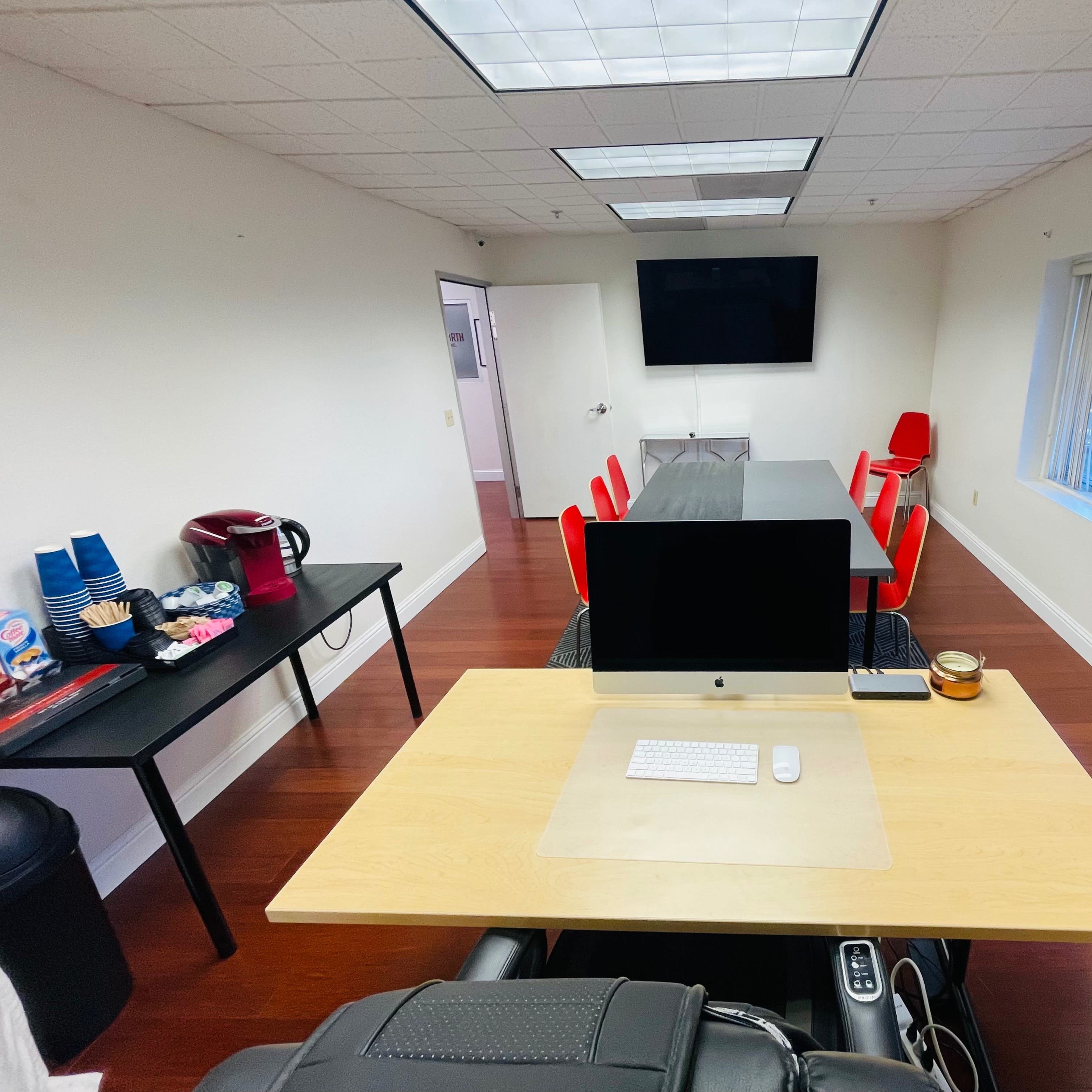 A modern office meeting room features a large conference table, a desktop computer on a wooden desk, and red chairs along with a kitchenette area.