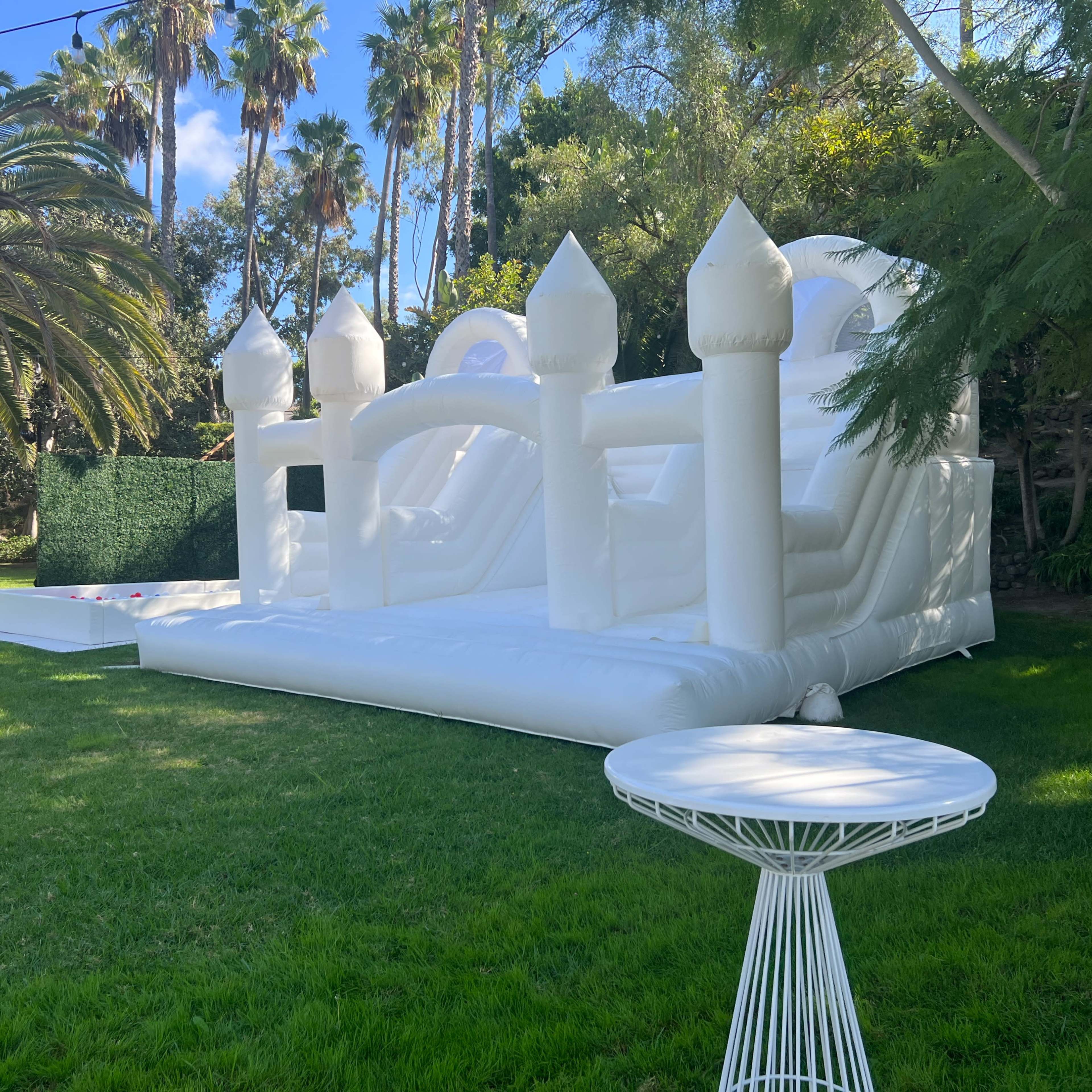 A large, white inflatable bounce house with turrets is set up on a grassy lawn surrounded by palm trees.