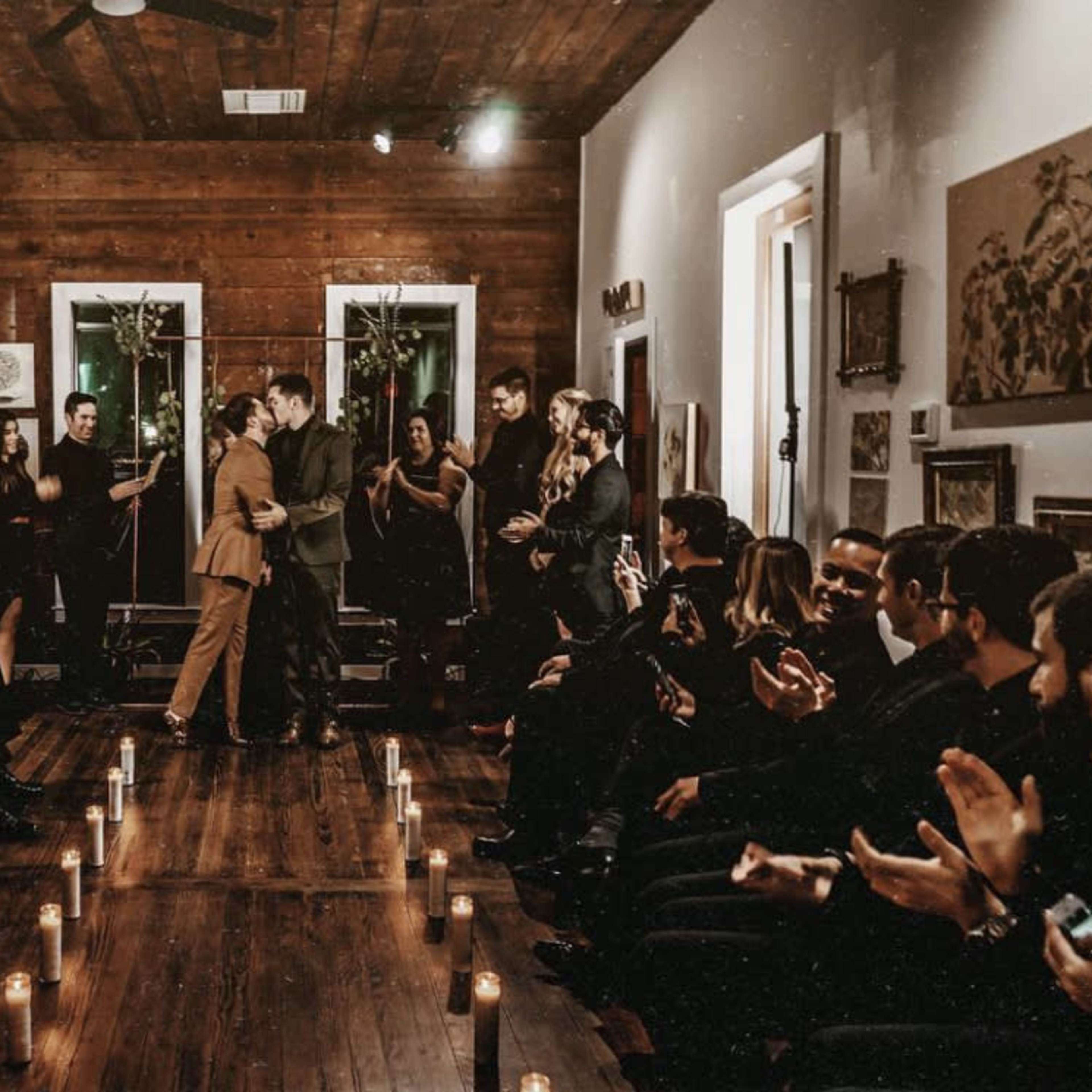 A couple stands facing each other and embracing at the front of a candlelit room filled with seated guests who are applauding.