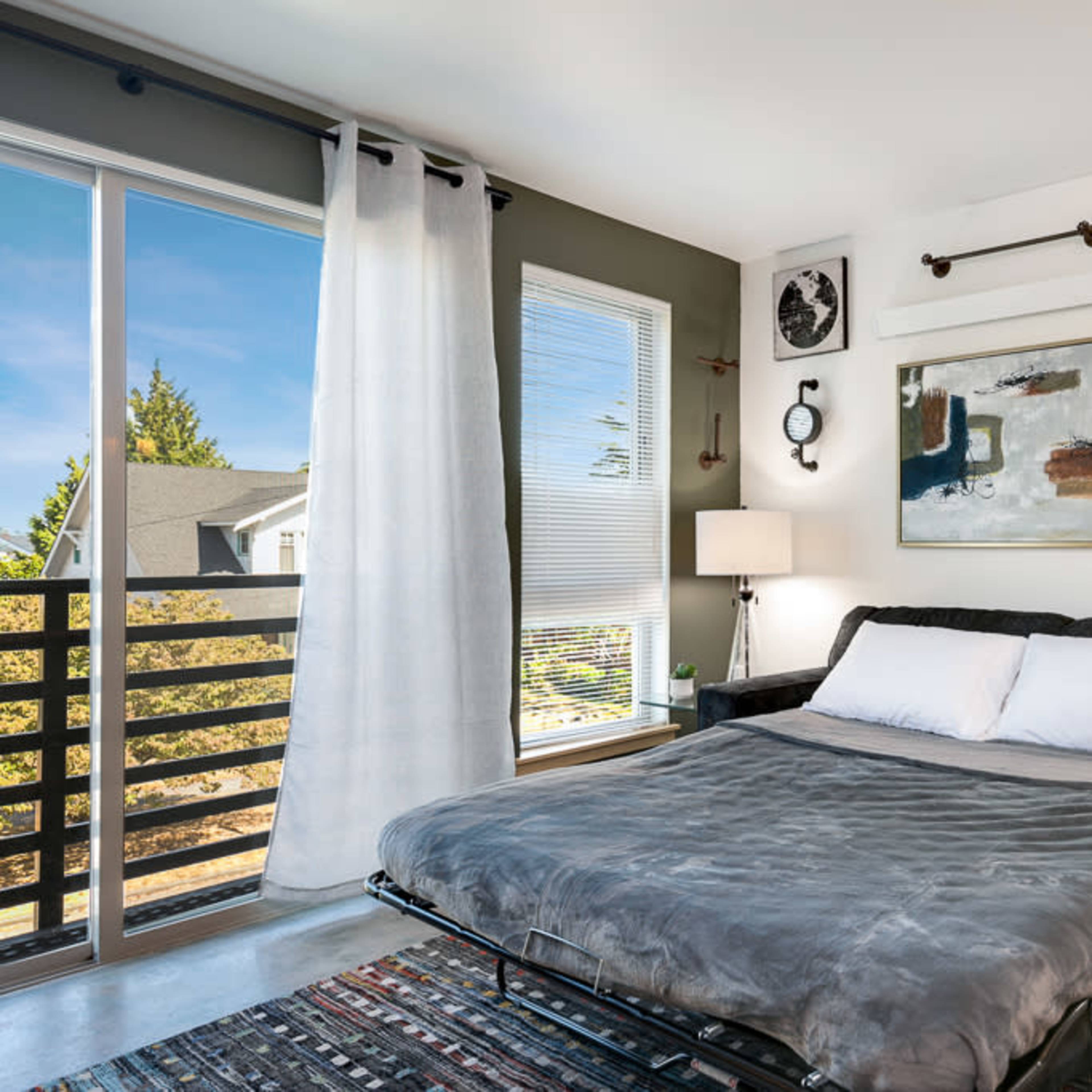A furnished bedroom features a fold-out sofa bed, a large window with sheer curtains, and modern artwork on the walls.
