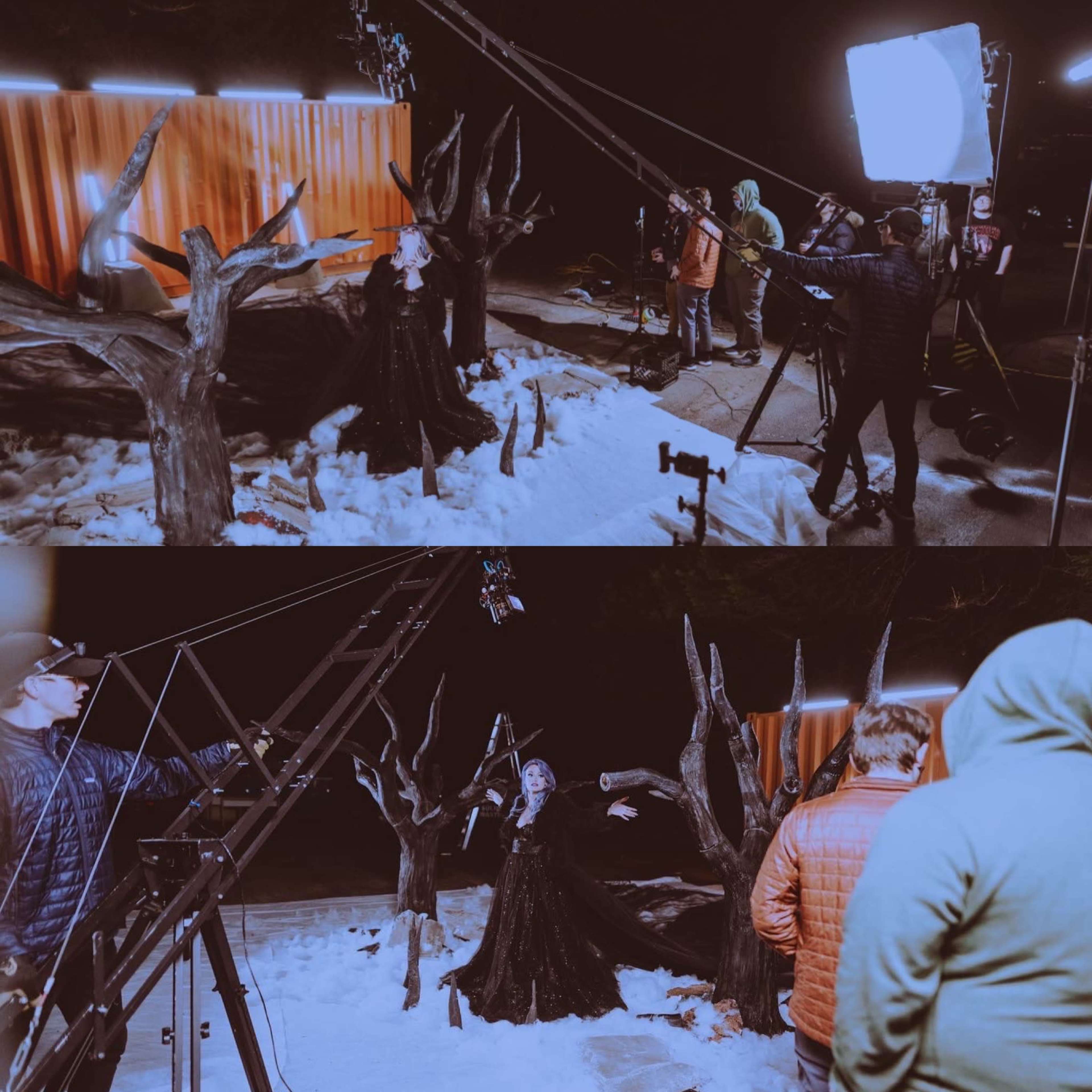 A film crew is filming a nighttime scene in the snow, featuring a woman dressed in black standing among trees with stark branches and bright lights illuminating the area.