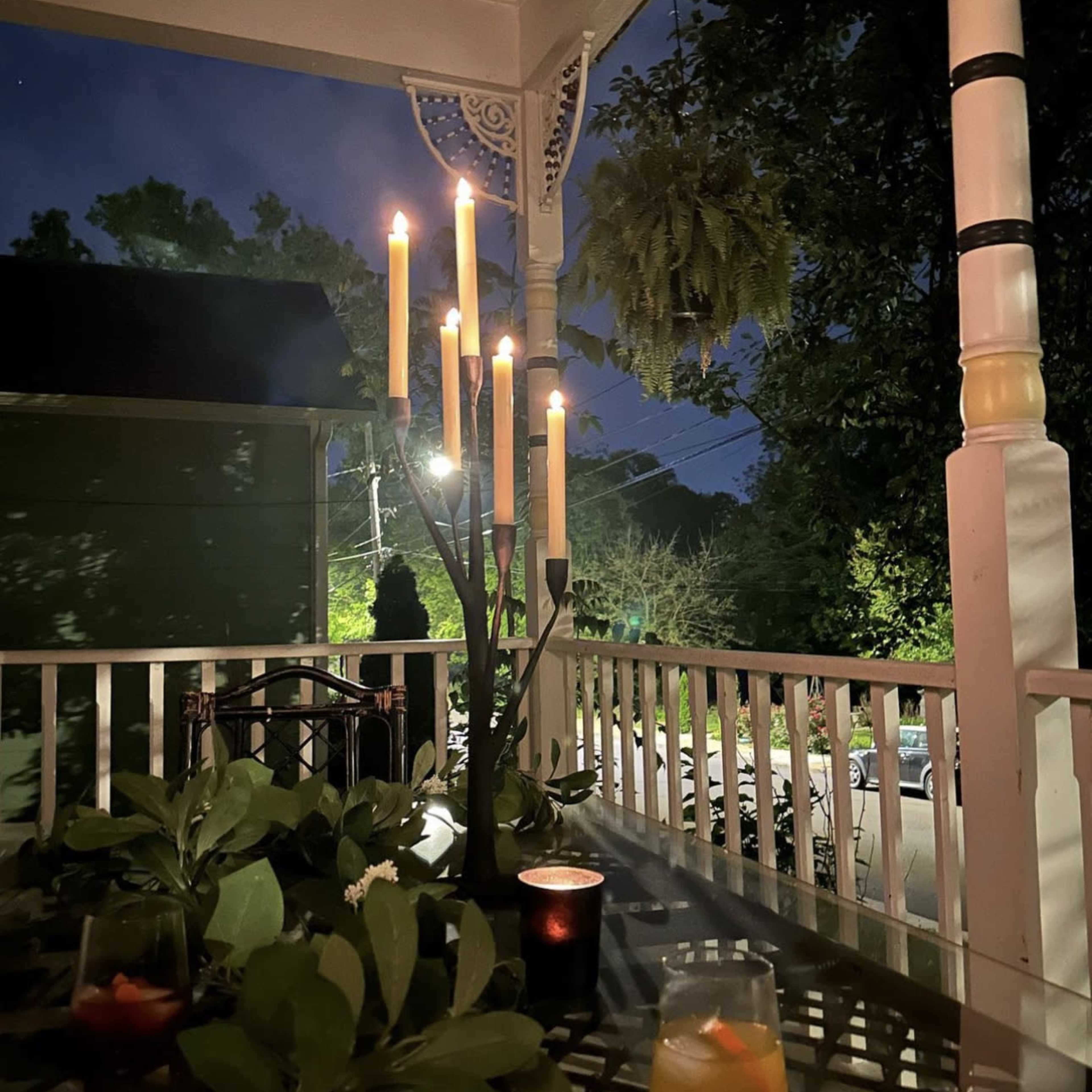 A candlelit table is set on a porch, surrounded by greenery, with a backdrop of evening sky and faintly lit trees.