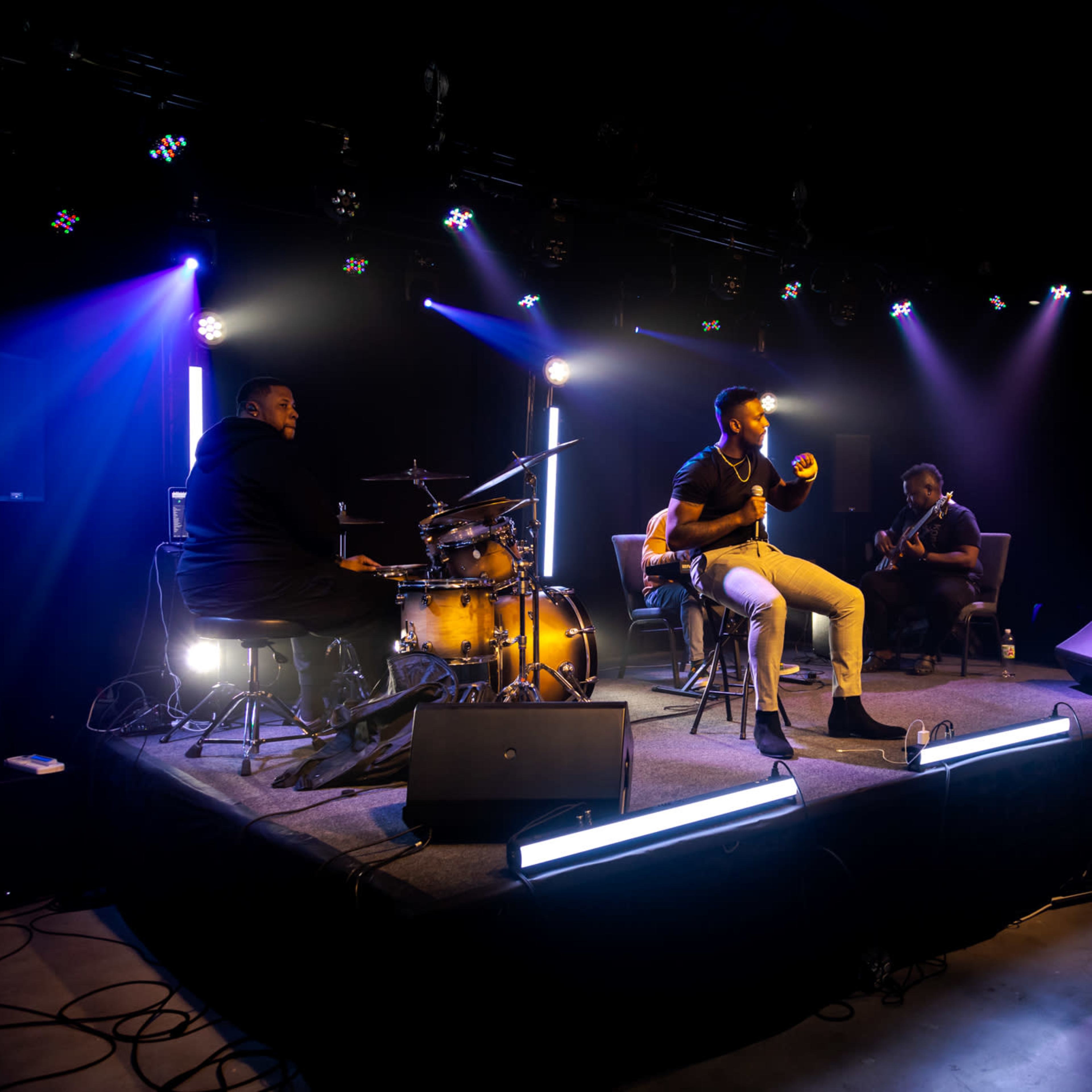 A musician performs on stage with a drummer and another guitarist in a dimly lit venue illuminated by stage lights.