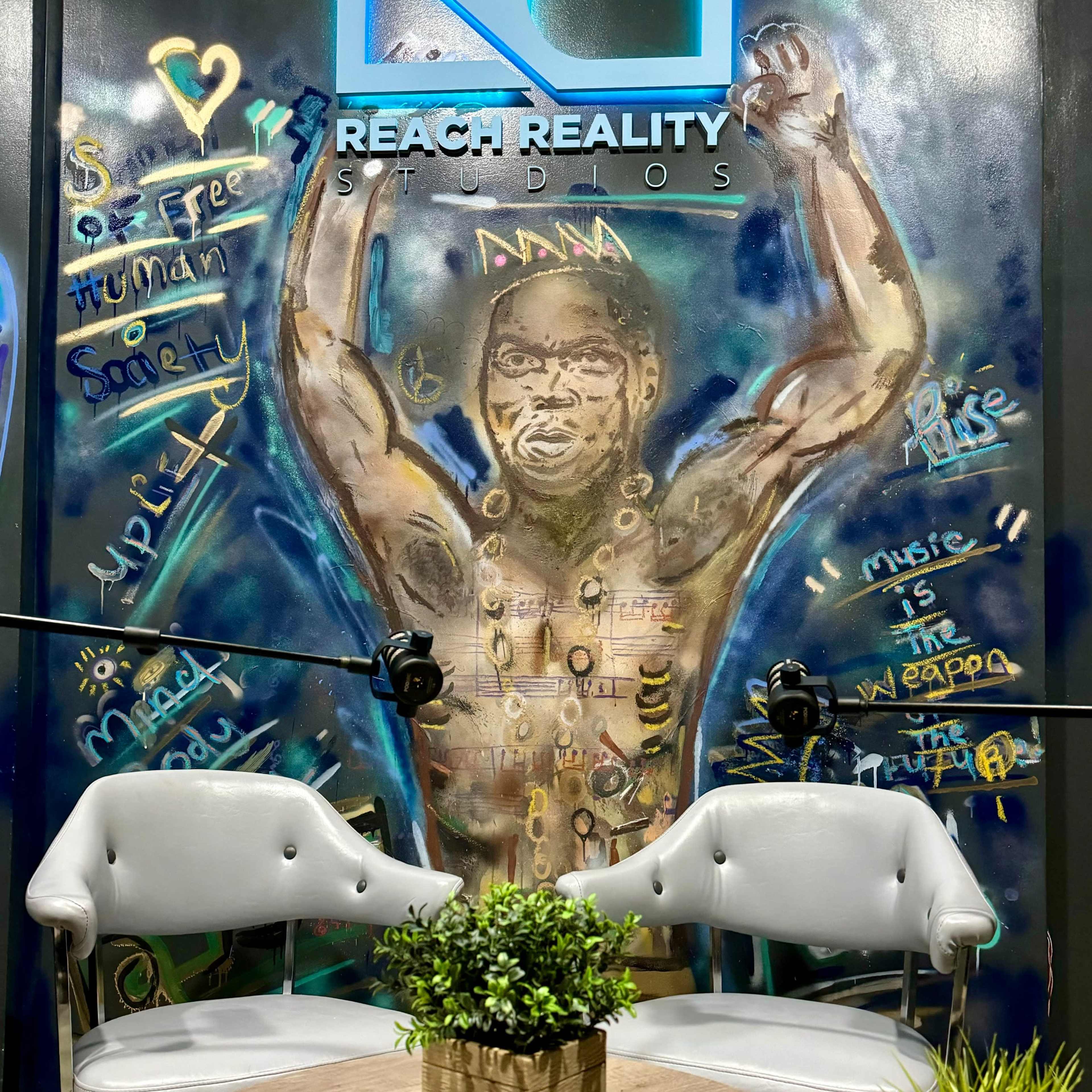 A colorful mural of a muscular man on a wall, with two gray chairs and a round wooden table featuring small potted plants in front of it.