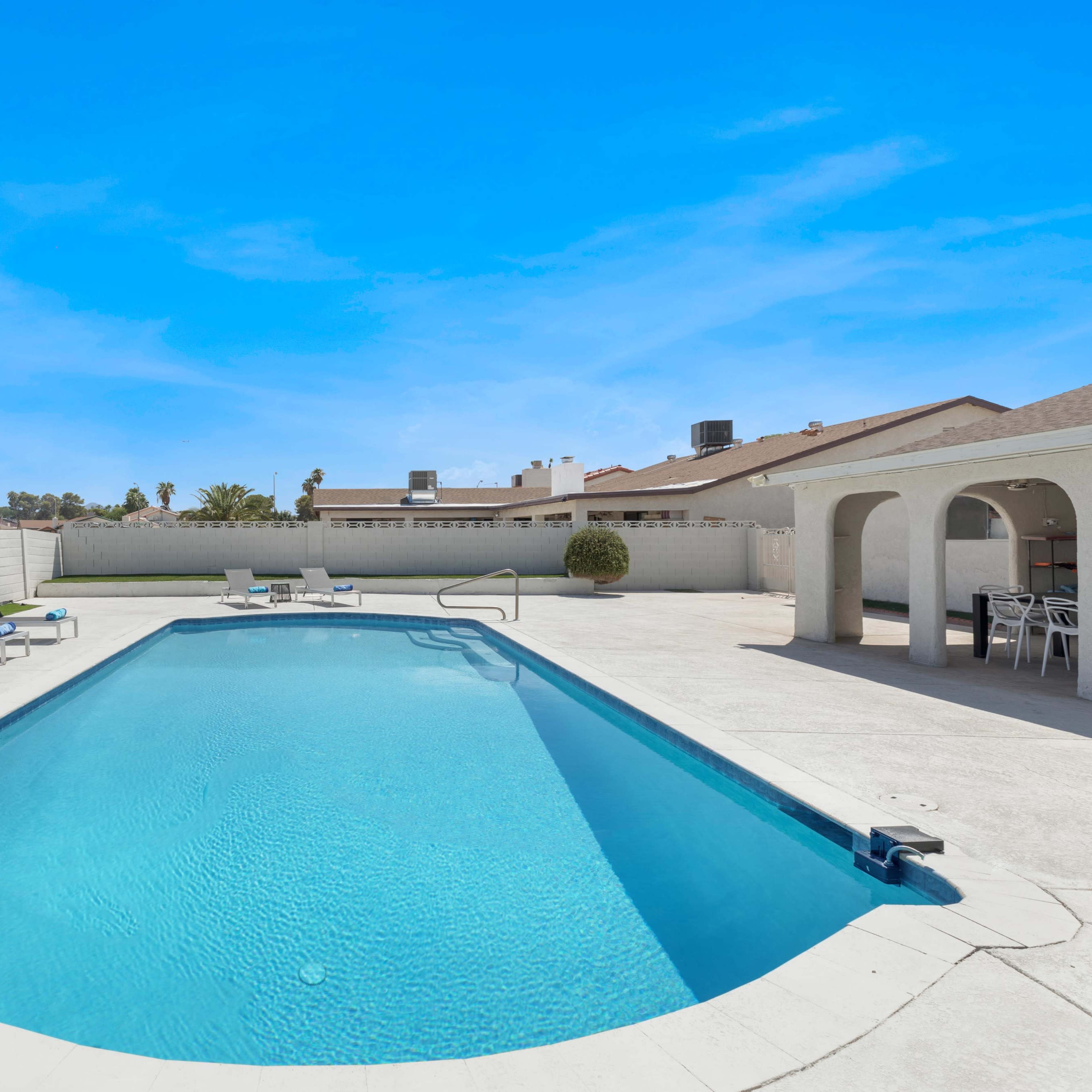 A rectangular swimming pool surrounded by lounge chairs, with a white wall in the background and a clear blue sky above.