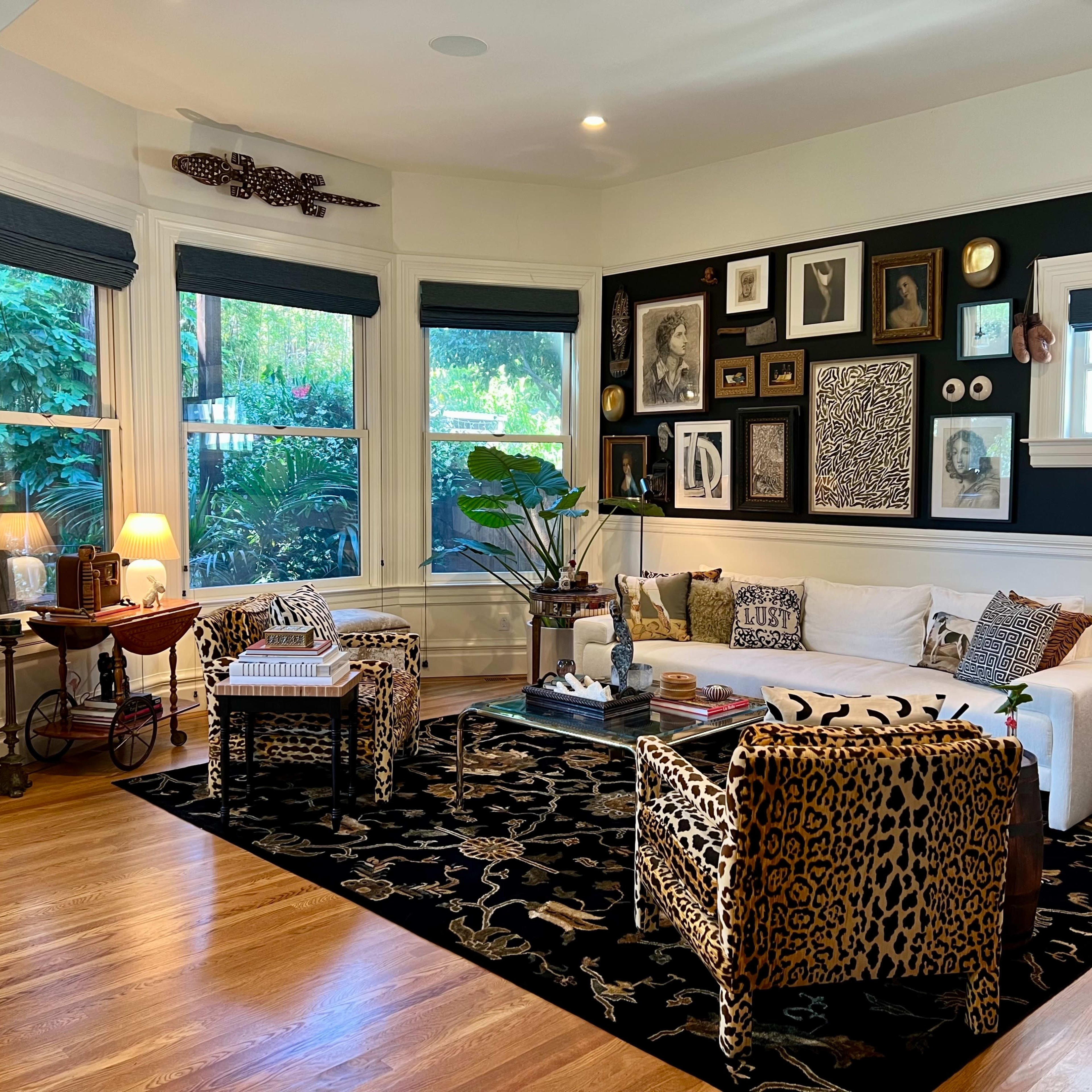 A stylish living room with a white sofa, leopard-print chairs, a black and gold patterned rug, and an eclectic wall display of framed art and photographs.