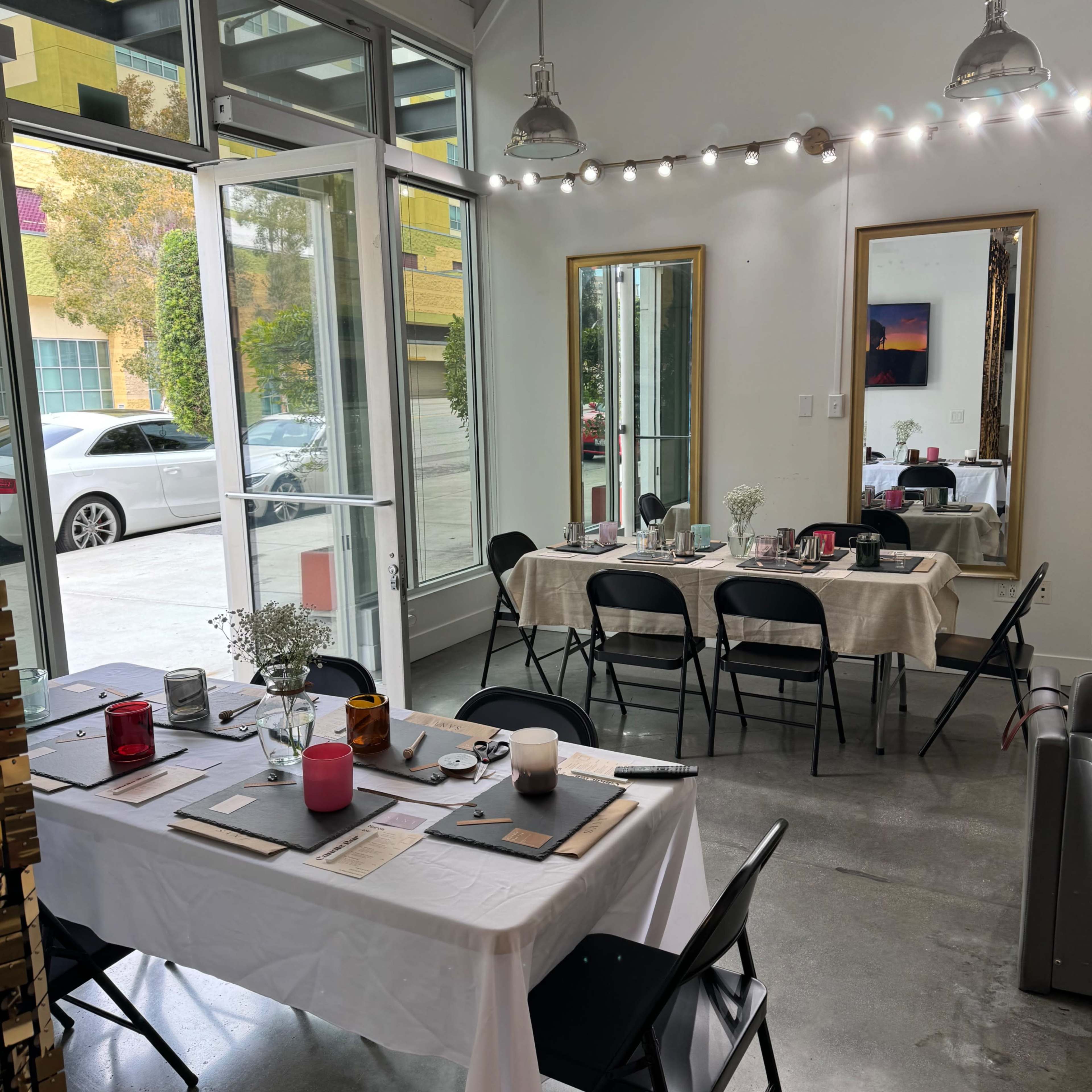 A modern, bright room set up for dining, featuring white tables with tablecloths, black chairs, and decorative mirrors, with large windows allowing natural light to enter.