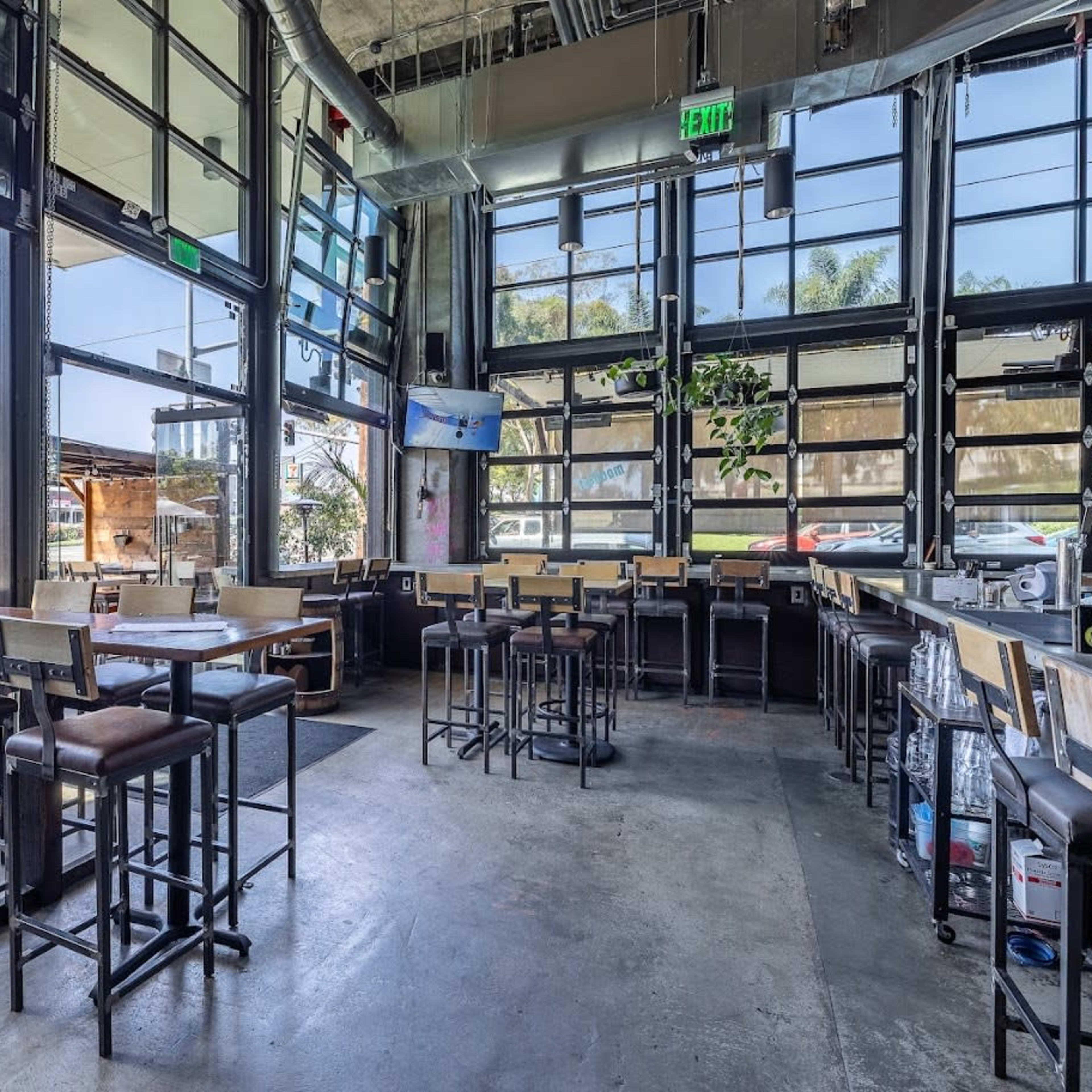The interior of a modern restaurant with large windows, high tables, and bar stools arranged around an open space.