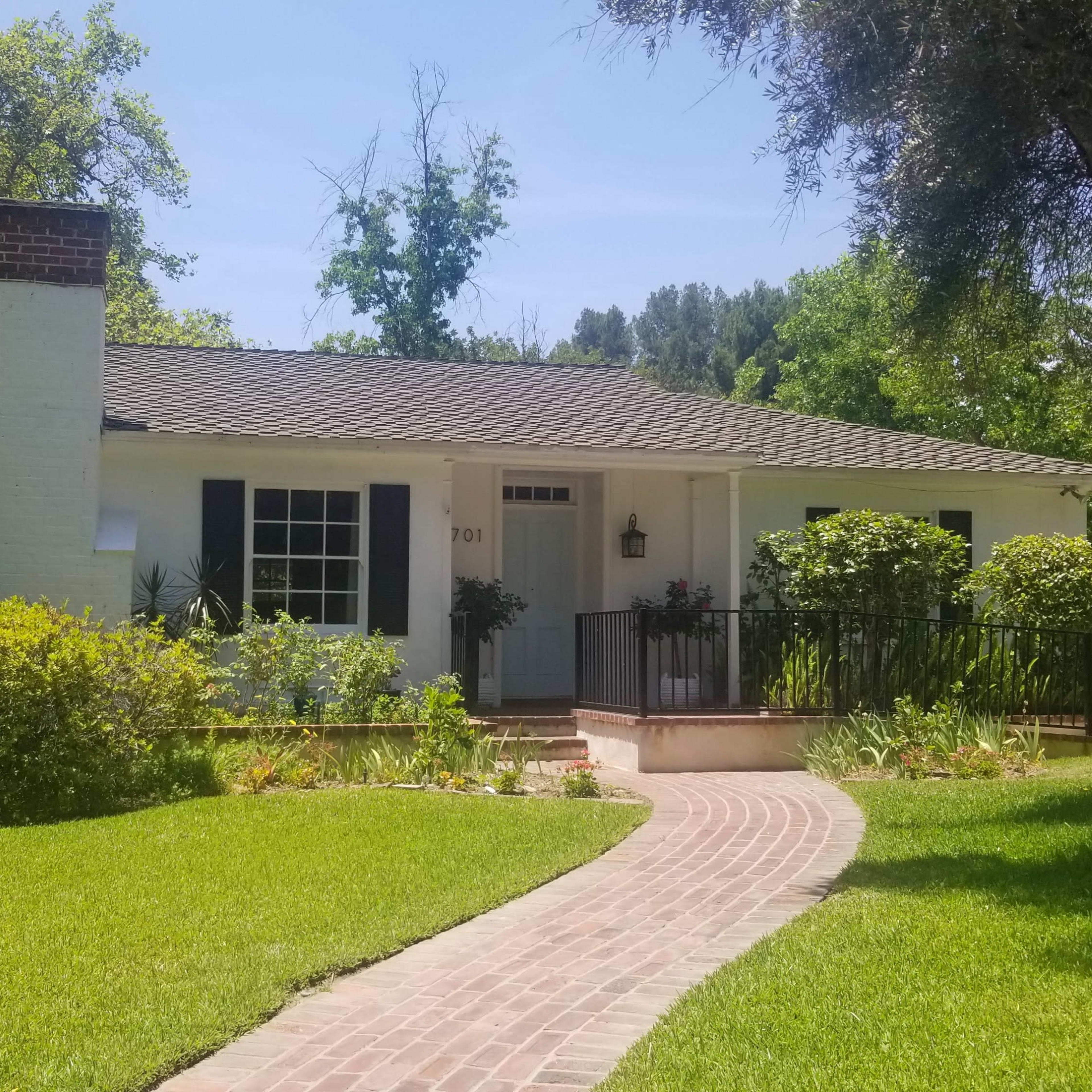 A single-story, white house with black shutters and a brick walkway is surrounded by lush greenery and trees.
