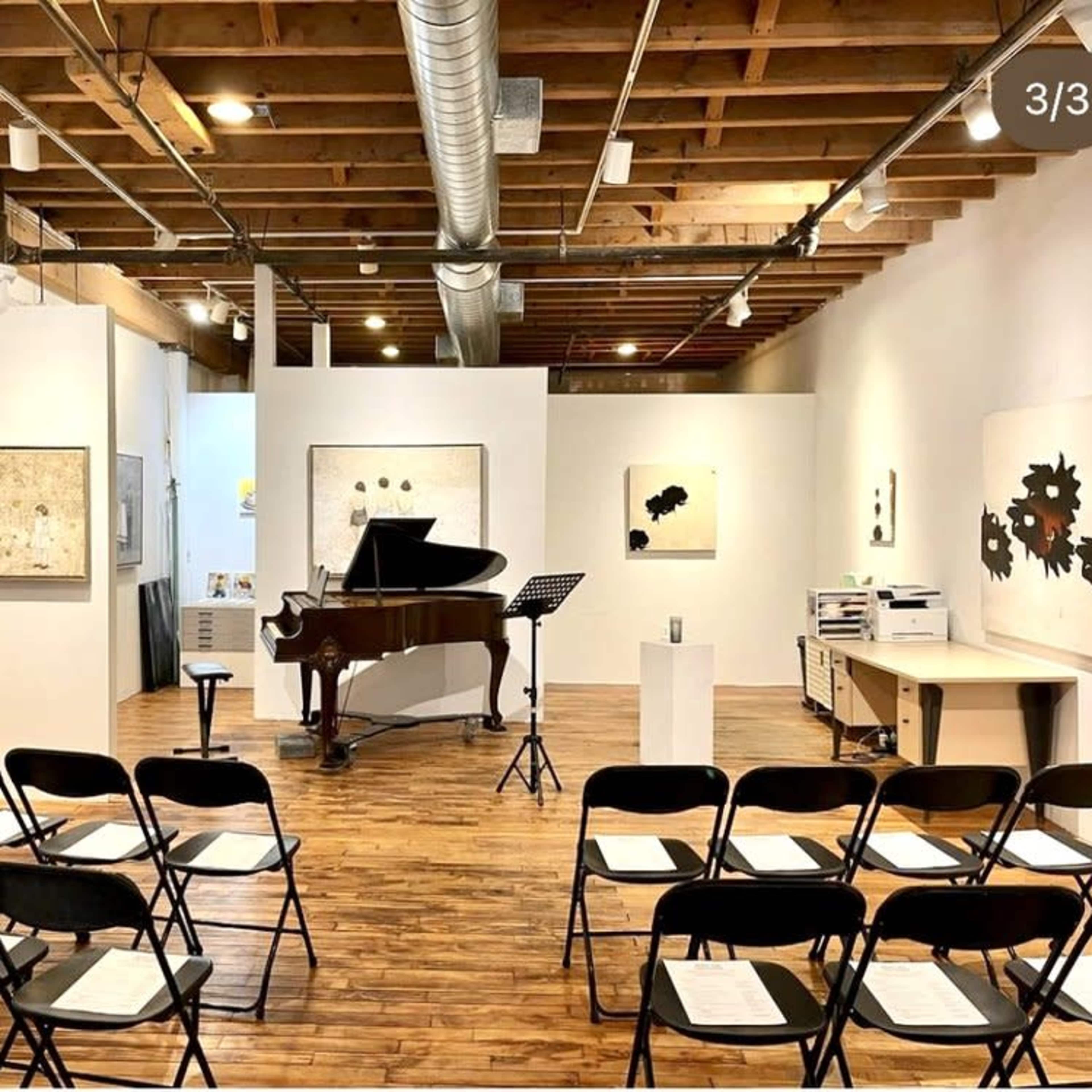 A music performance space with a grand piano in the center, surrounded by rows of black chairs and art displayed on the walls.