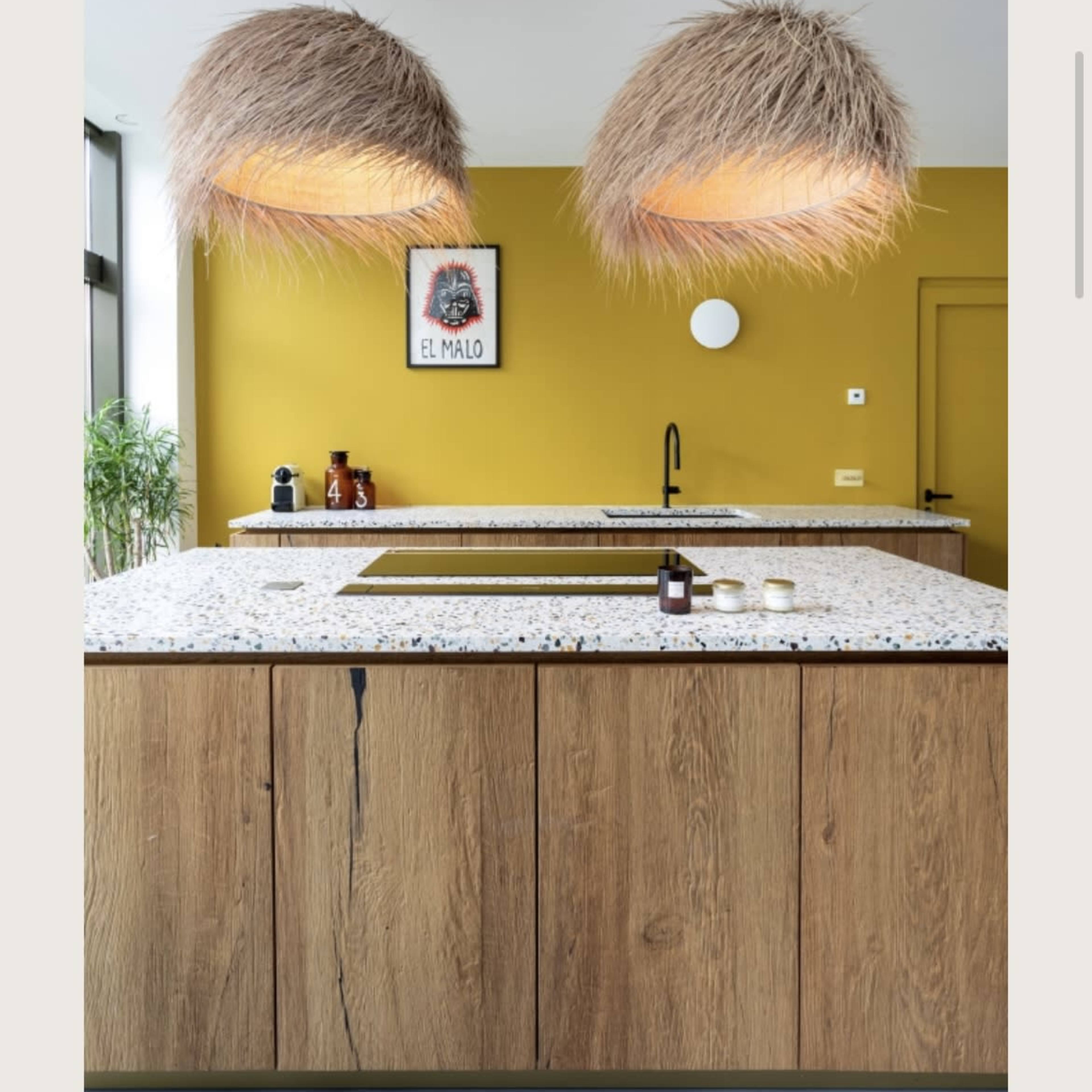 A modern kitchen with a yellow wall, two large pendant lights made of straw, and a central island with a speckled countertop.