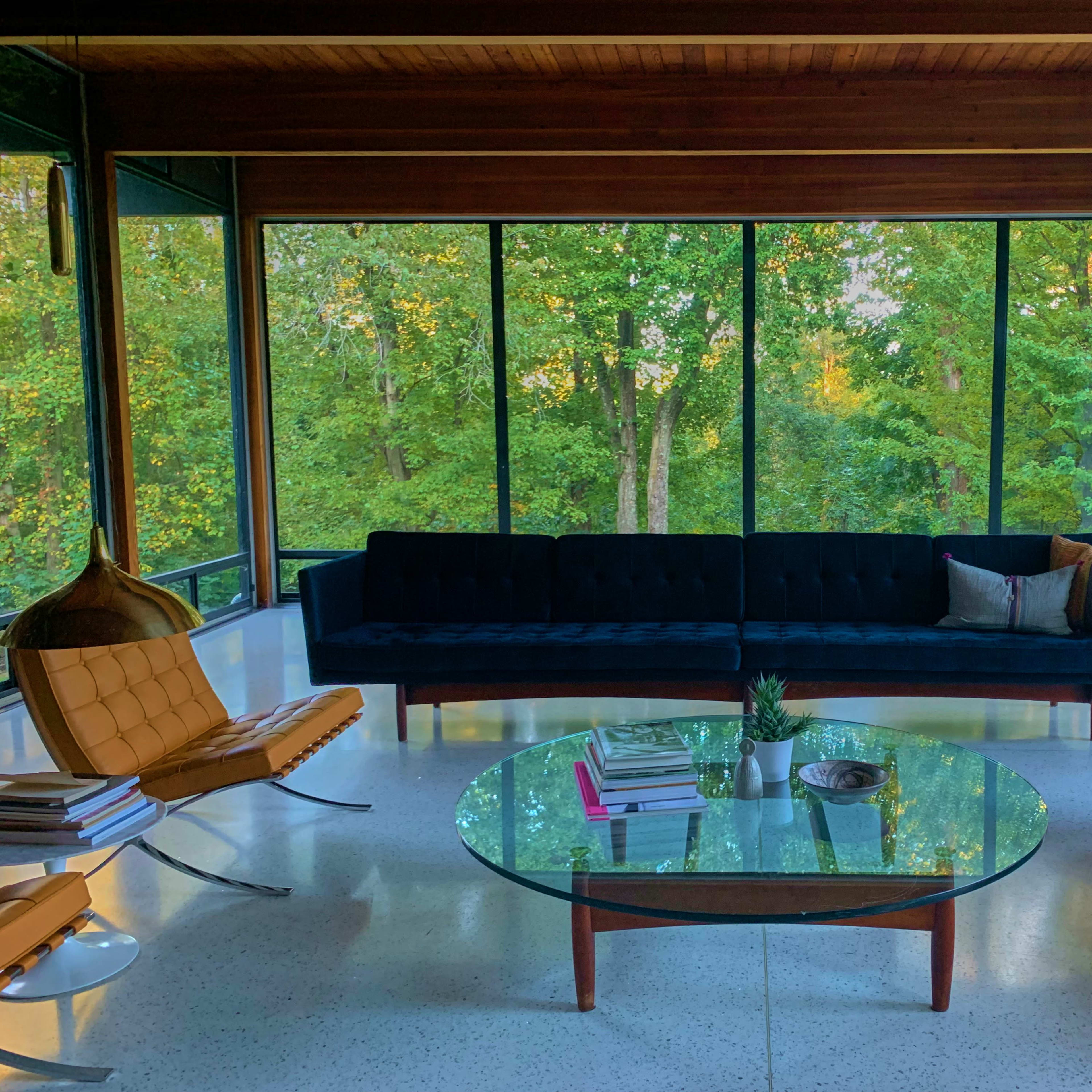 A modern living room with large windows overlooking a green wooded area, featuring a dark sofa, a glass coffee table, and two stylish chairs.