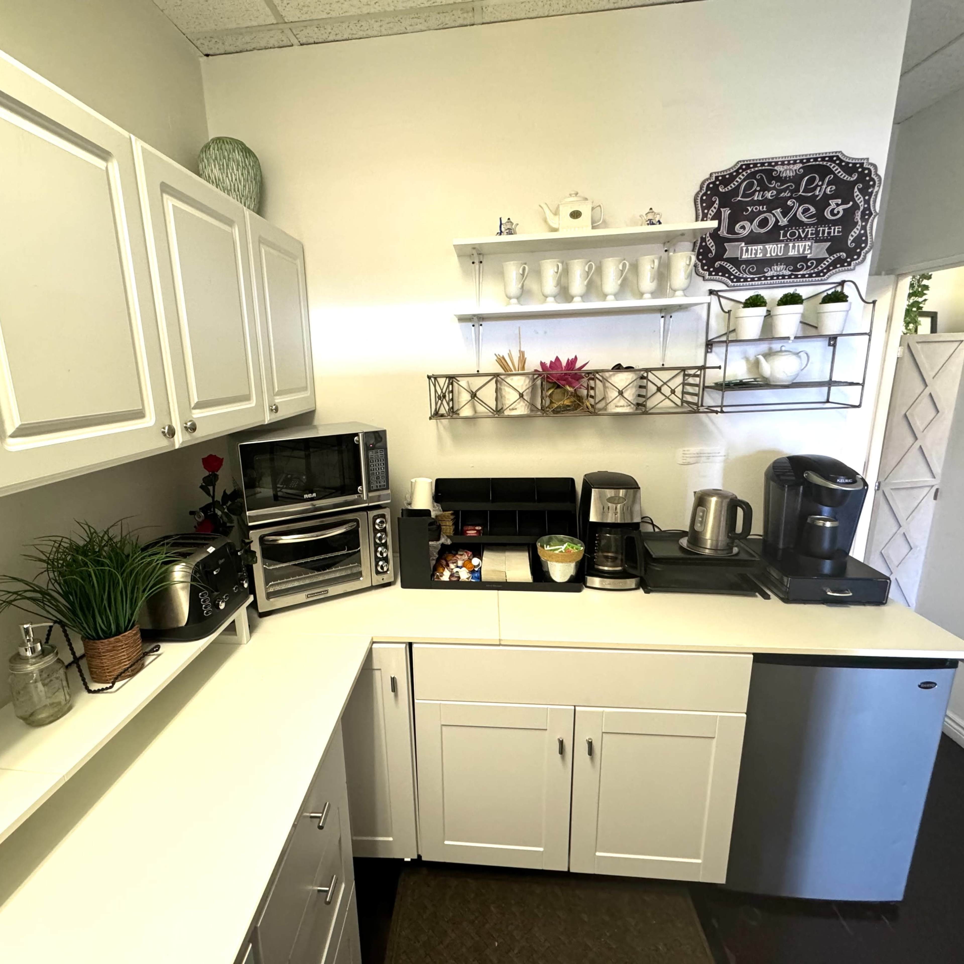 A well-organized kitchen corner with white cabinets, a countertop, various appliances, and decorative items on shelves.