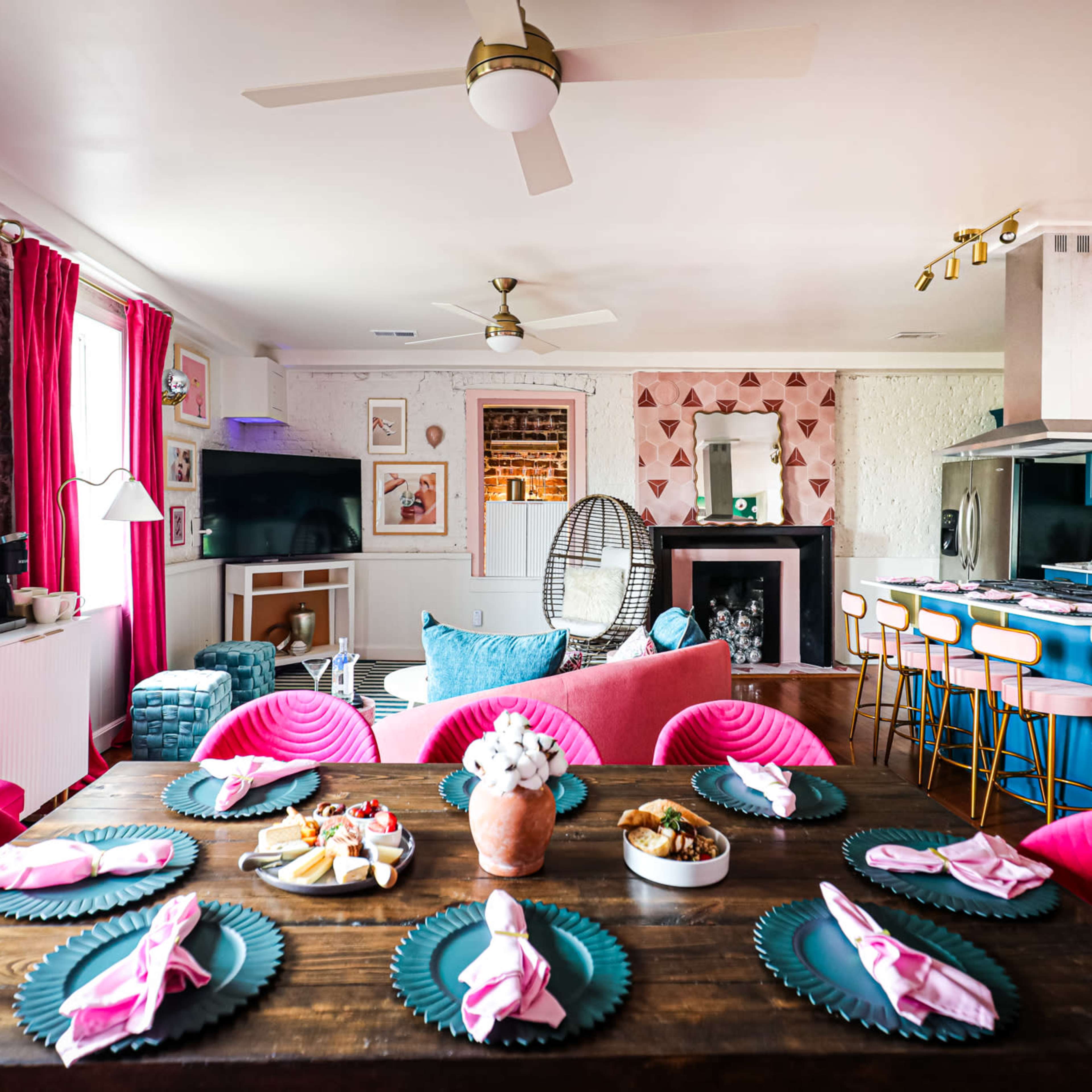 A vibrant living space with a dining table set for a meal, pink chairs, a fireplace, and a modern kitchen area in the background.