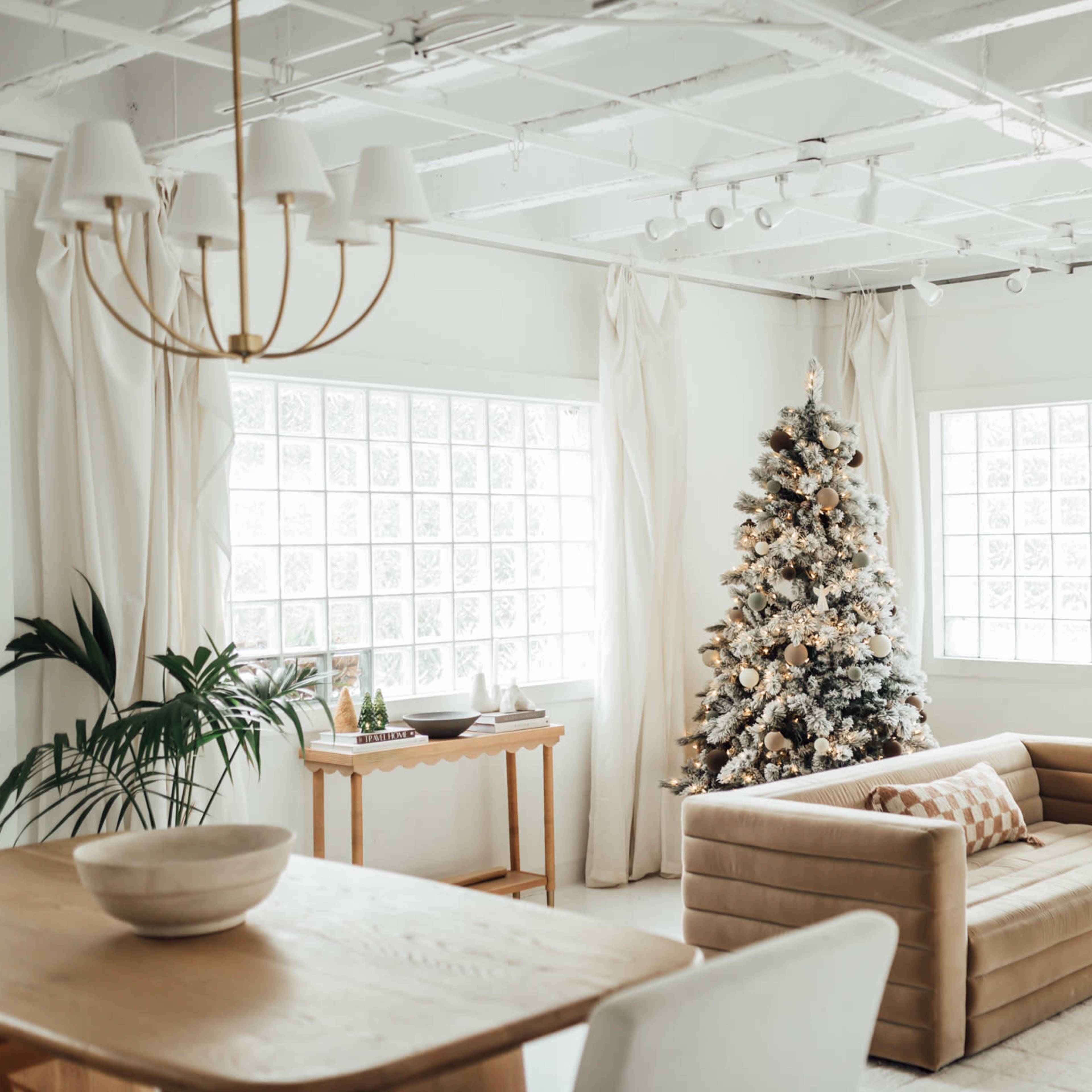 A cozy living room features a decorated Christmas tree, large windows, and a beige sofa, with a dining table and greenery accents nearby.