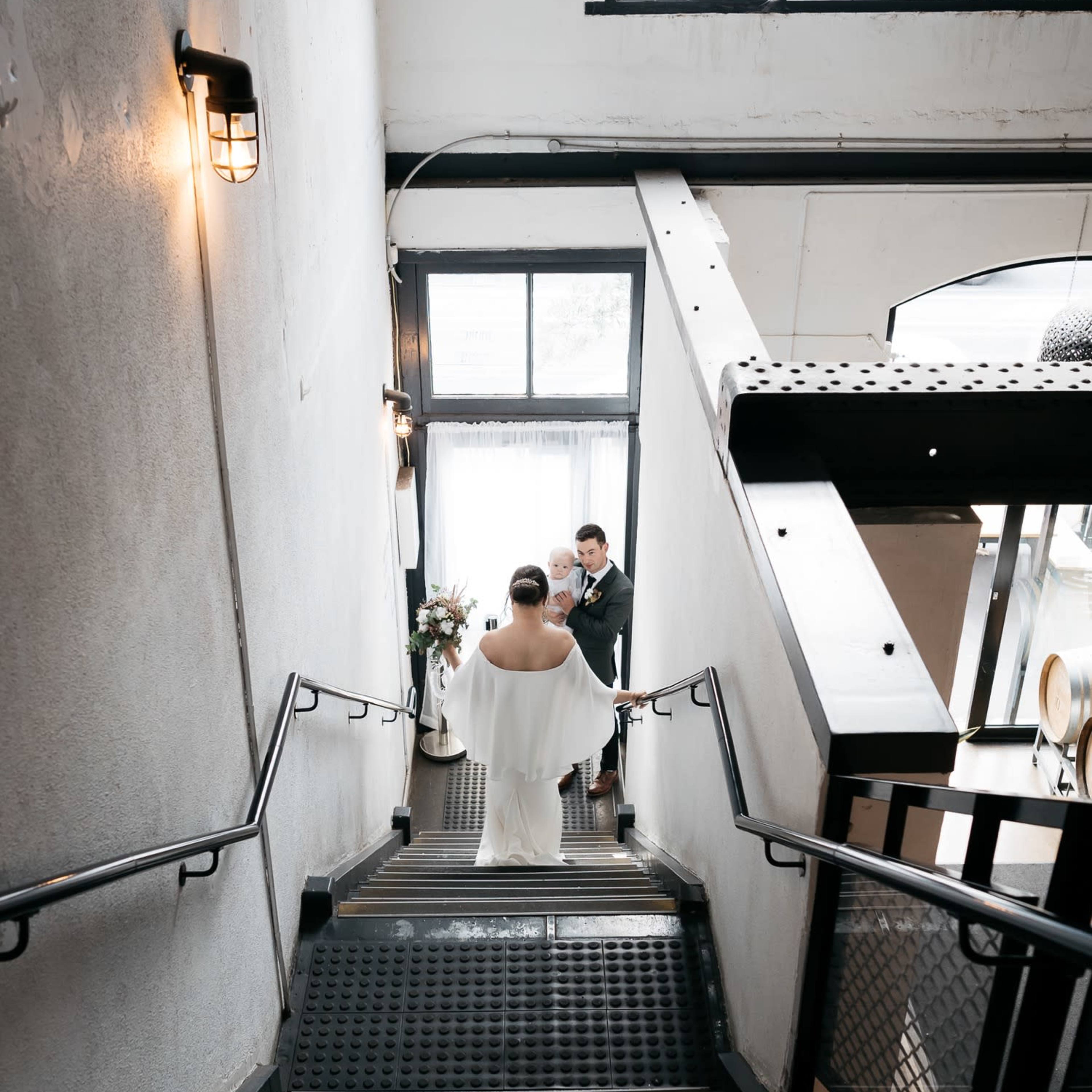 A couple stands on a staircase, with one partner descending and the other waiting at the bottom, surrounded by industrial-style decor and large windows.