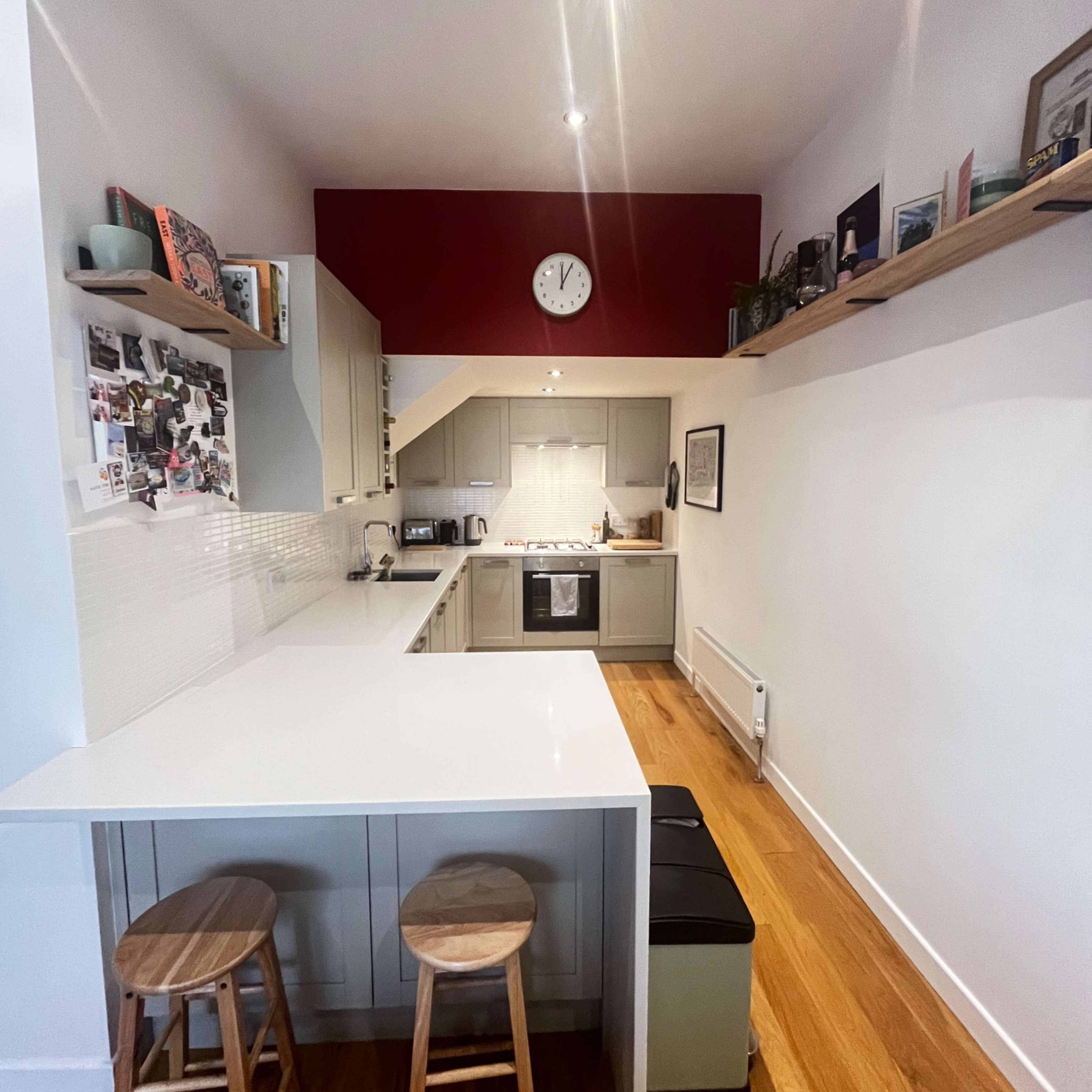 A modern kitchen features a white countertop, wooden stools, and a red accent wall with a clock.