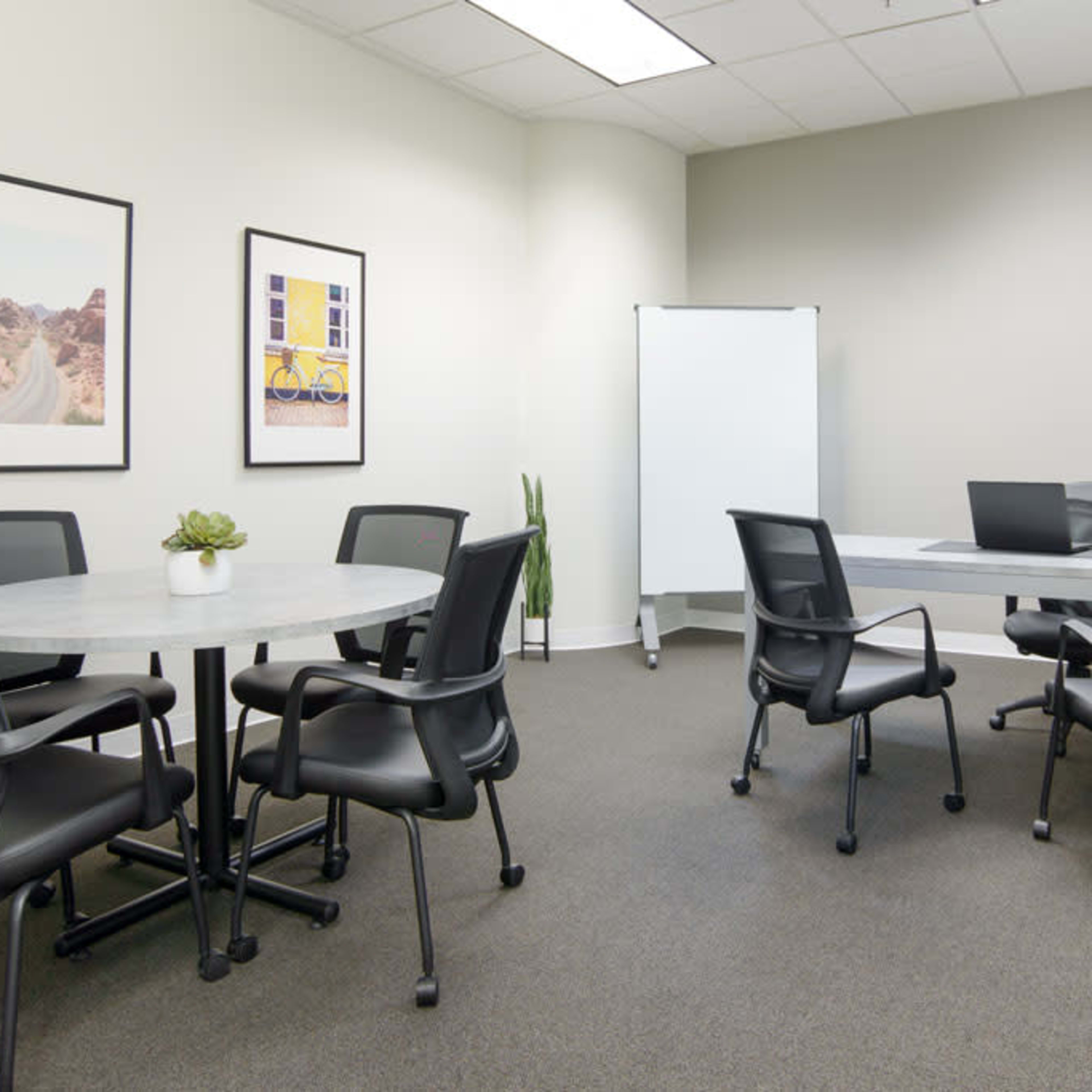 A minimalistic meeting room with a round table, six black chairs, a whiteboard, and a laptop on a rectangular table.