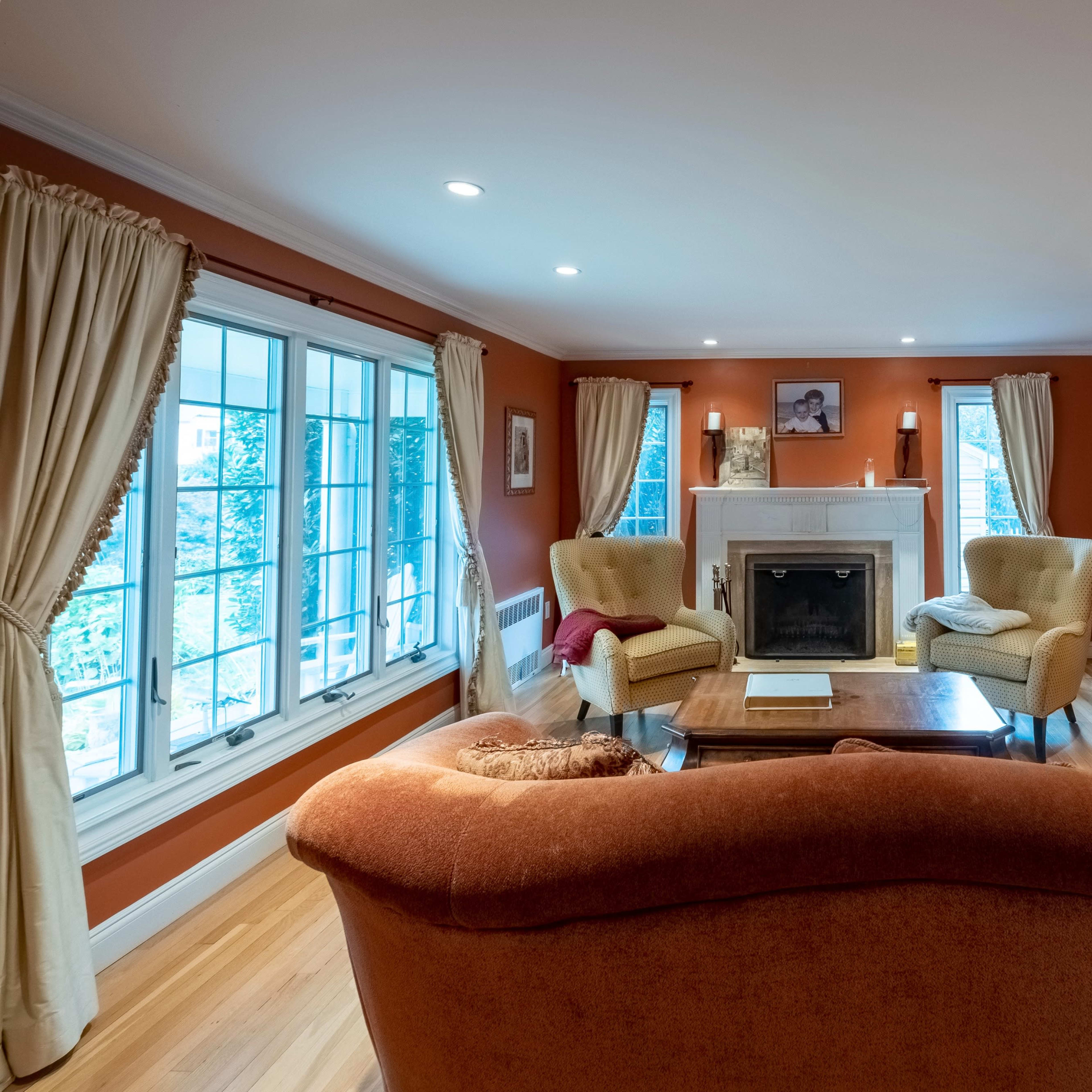 A cozy living room features an orange wall, a fireplace, two armchairs, and large windows allowing natural light.