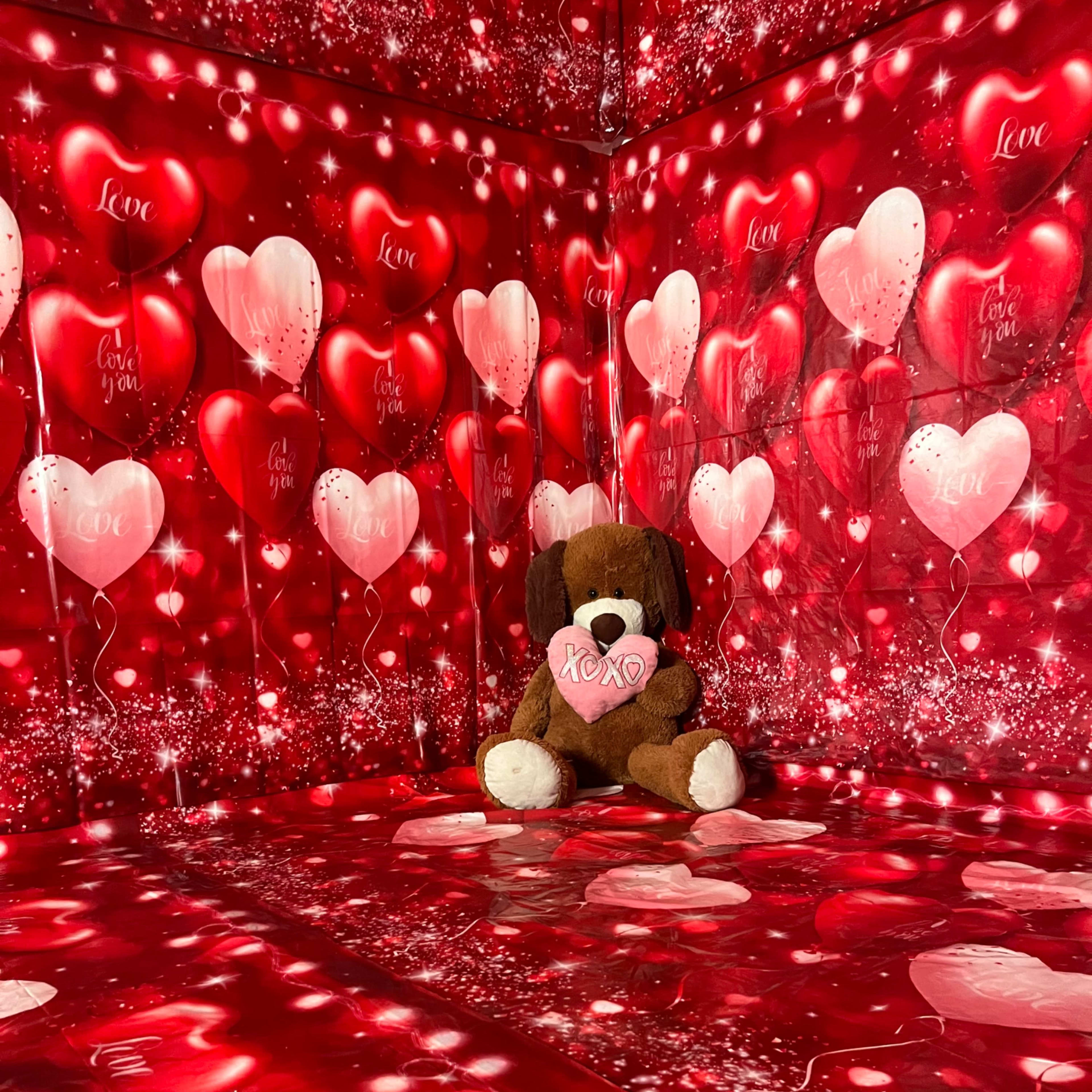 A stuffed dog sits in a corner of a room decorated with red and pink heart-themed wallpaper and floor covering.