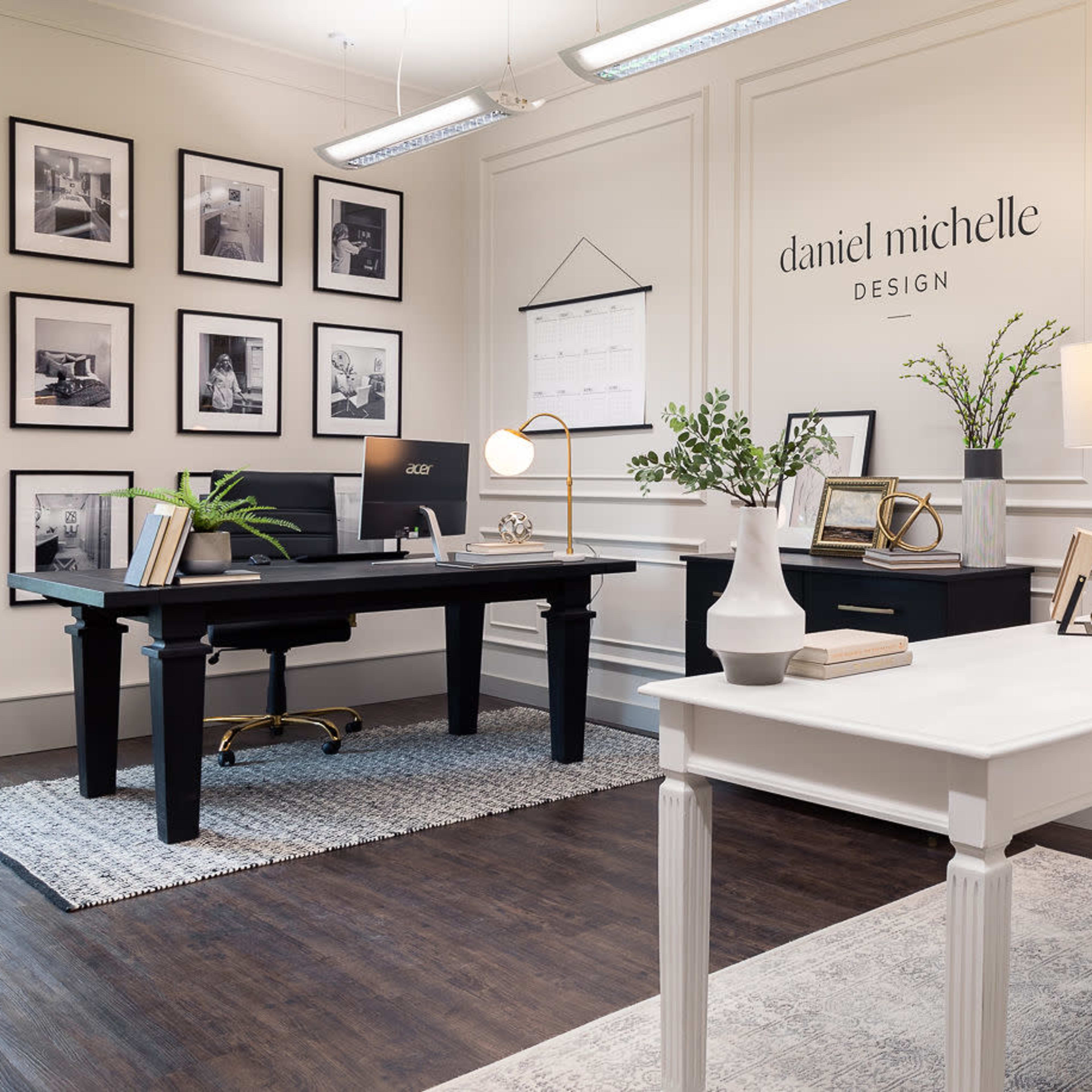 A modern office space with a black desk on the left, a white desk with a gold-accented chair on the right, and a gallery wall of framed photos in a well-lit room.