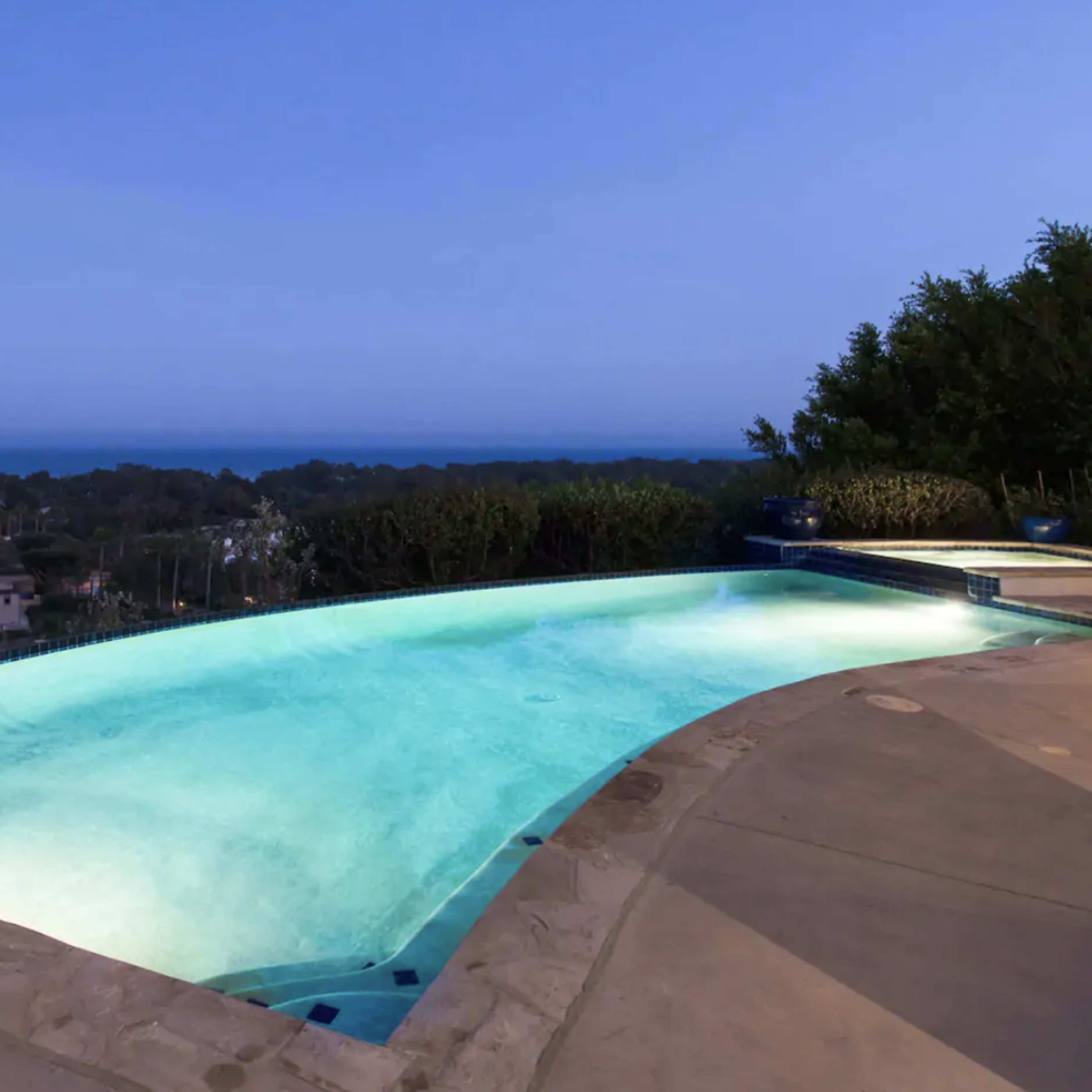 A glowing swimming pool overlooks a coastal landscape at dusk.