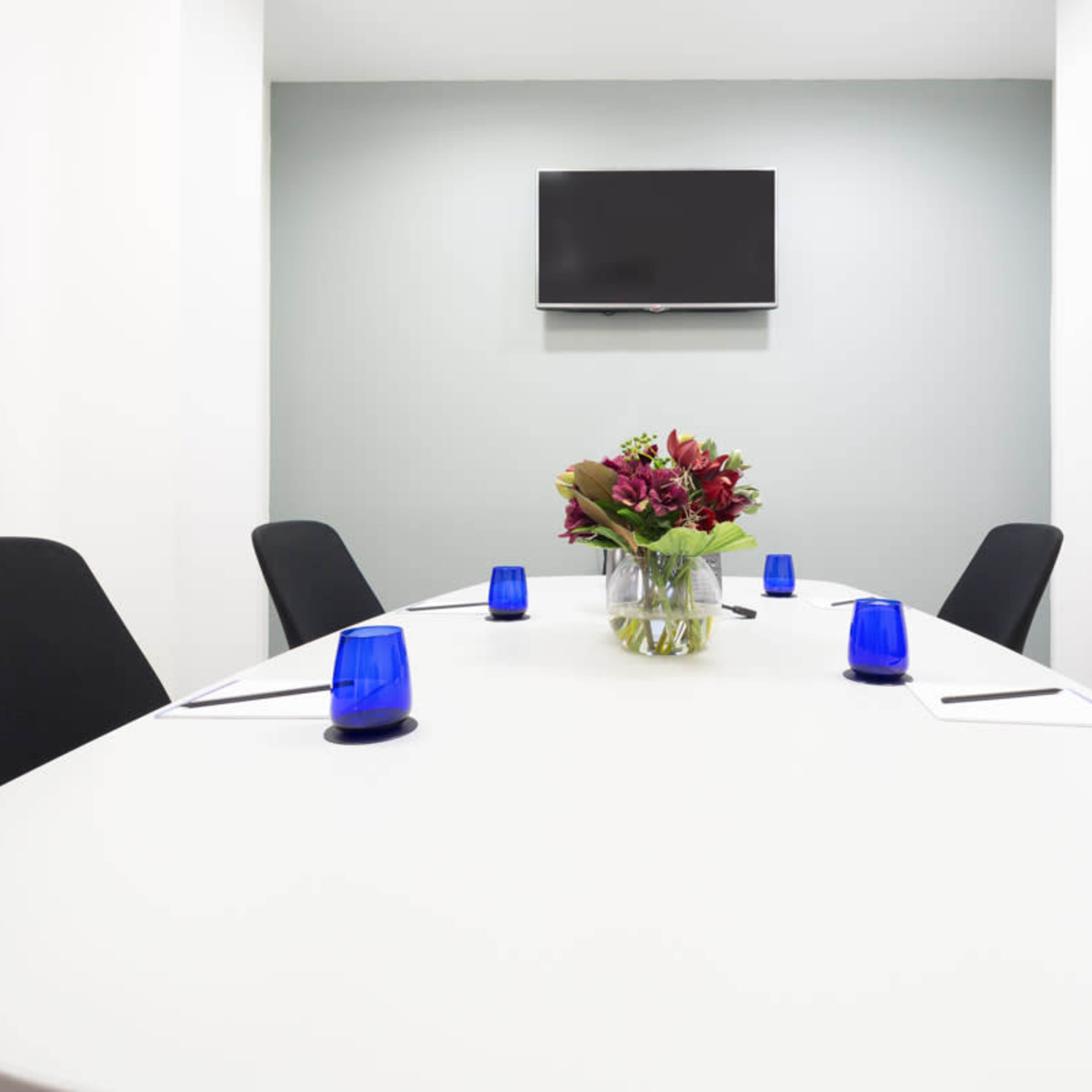 A conference room features a white oval table with a floral centerpiece, surrounded by black chairs, and a wall-mounted television.